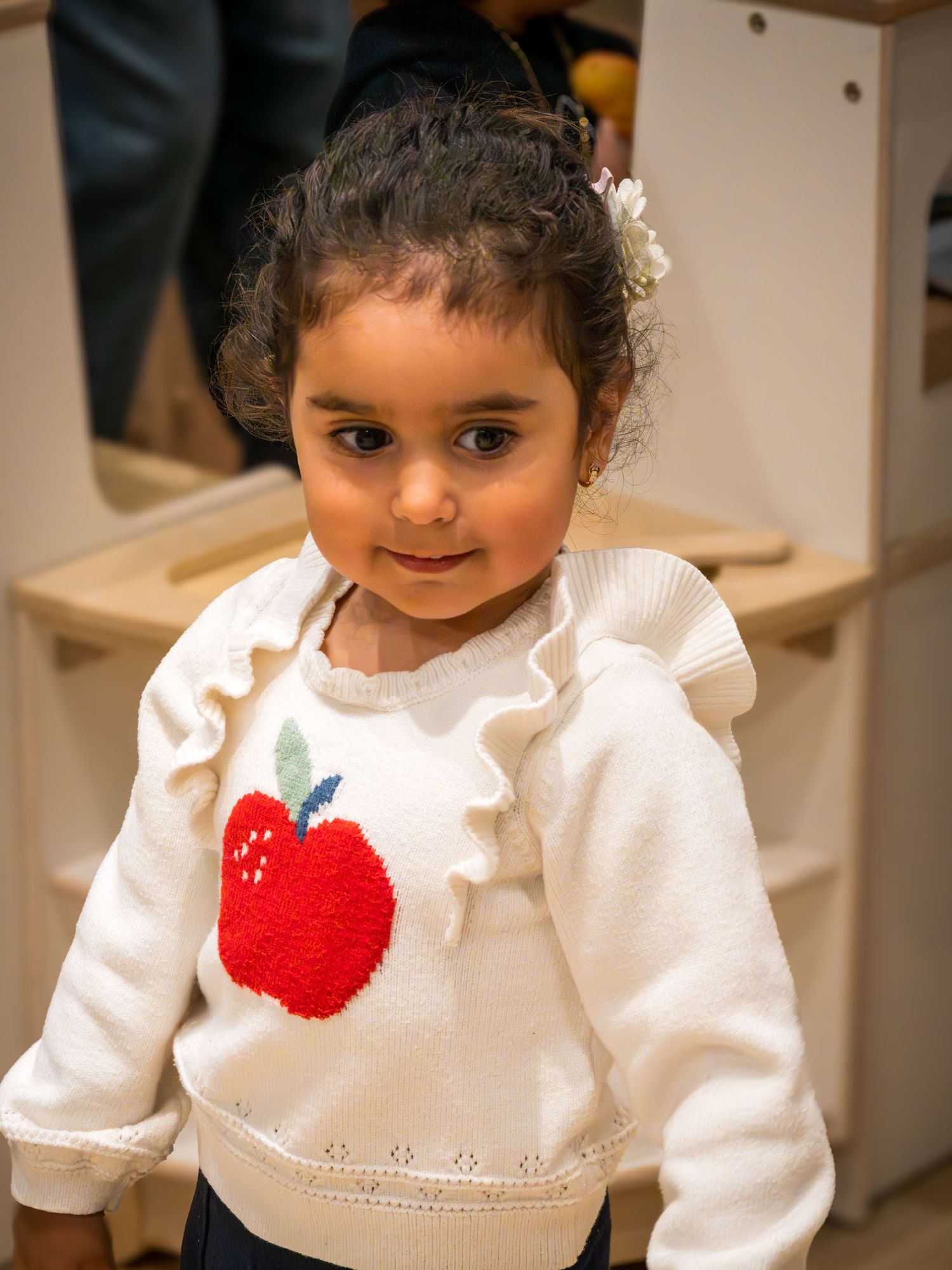 Cute toddler girl in white apple print ruffle sweater with hair accessory looking at camera.
