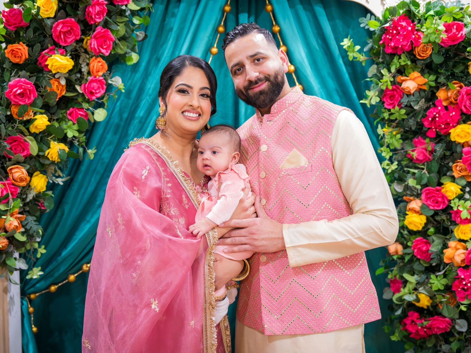 Happy South Asian couple in matching pink traditional outfits holding infant at colorful floral backdrop.