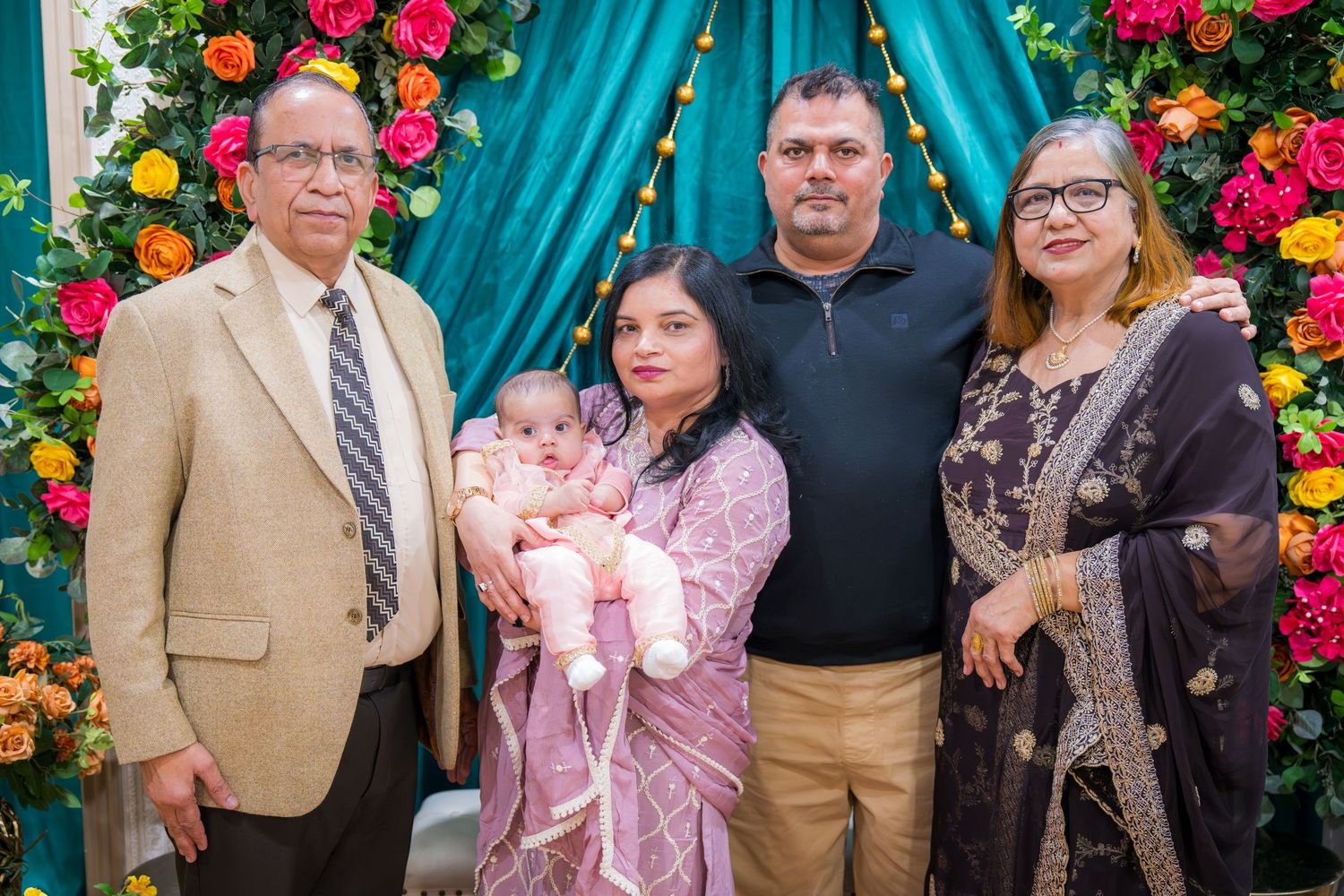 South Asian family of five with newborn baby posing before vibrant floral ceremony backdrop.