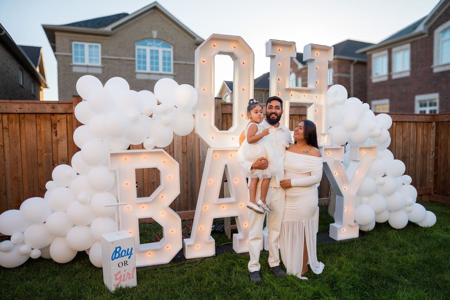 Family poses at outdoor gender reveal party with lit OH BABY marquee letters and white balloon arch.