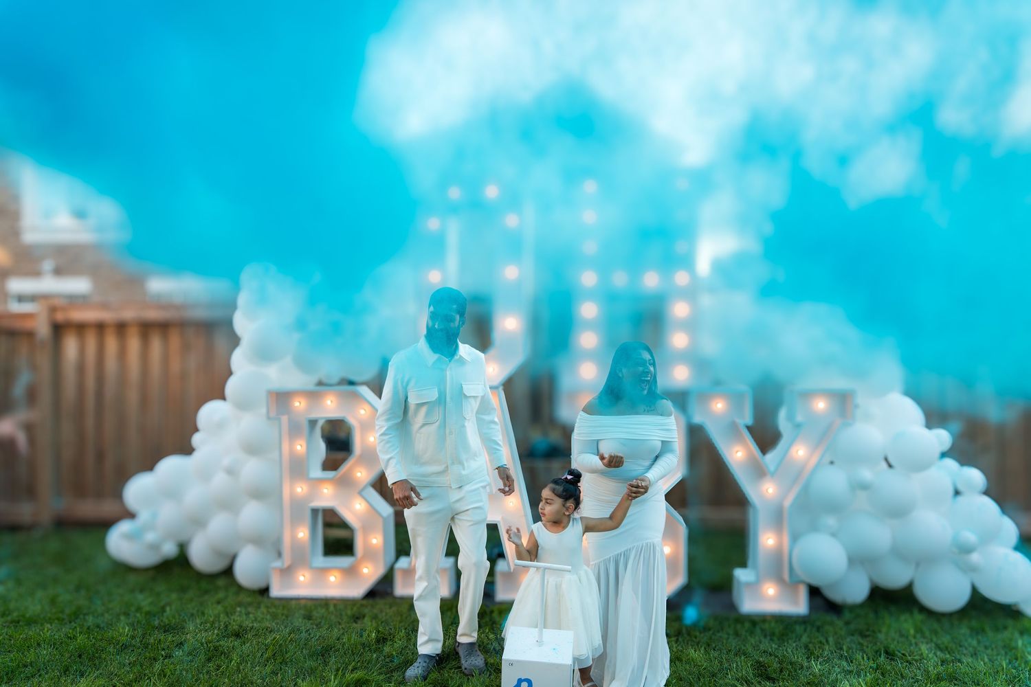 Couple with child standing before illuminated BABY marquee letters surrounded by blue gender reveal smoke.