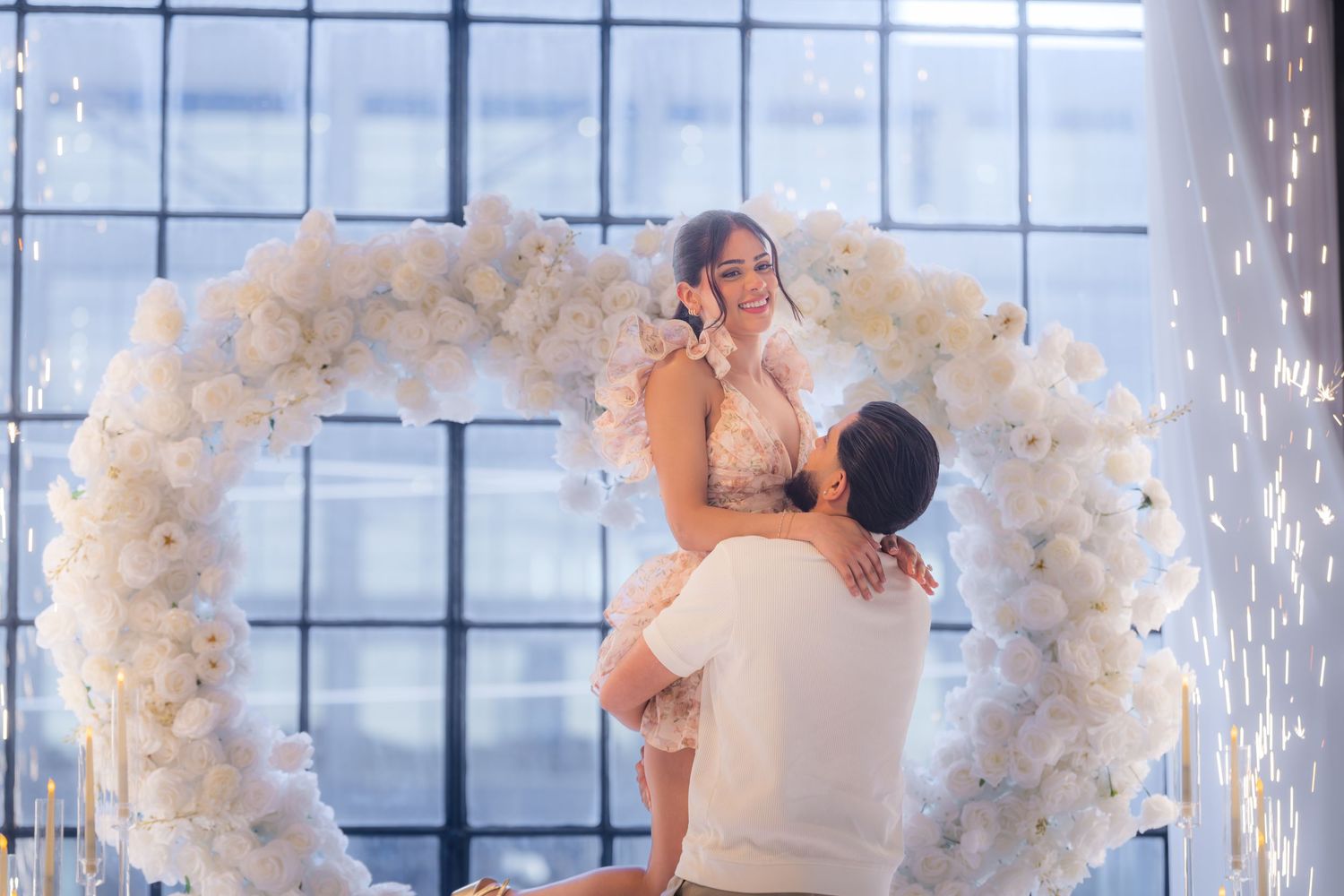Emotional marriage proposal moment with man lifting woman beneath white floral heart arch with sparklers.