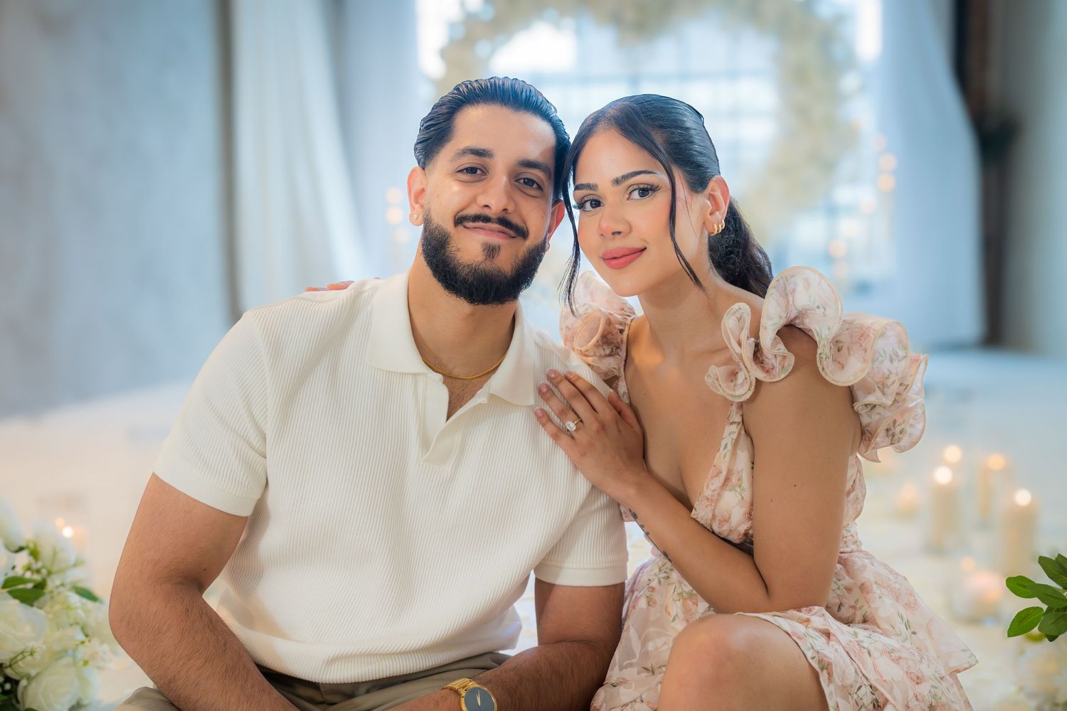 Smiling engaged couple posing together with white floral heart arch in background.