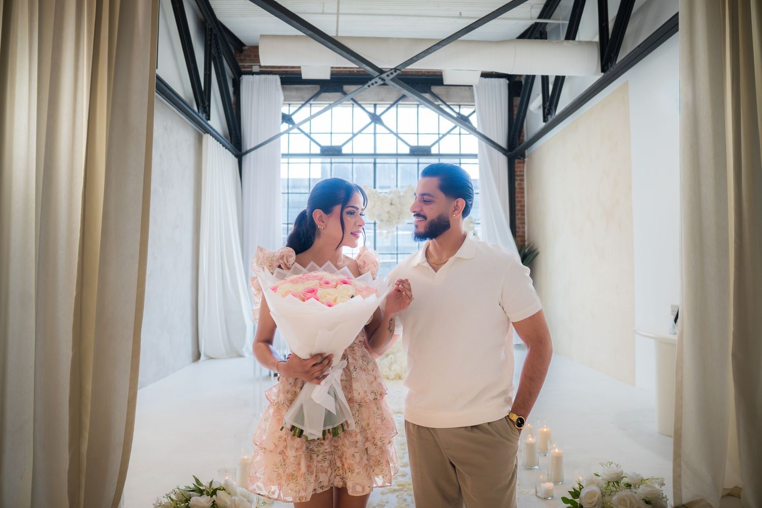 Happy couple sharing a romantic moment indoors, woman holding a large bouquet of pink roses during a marriage proposal.