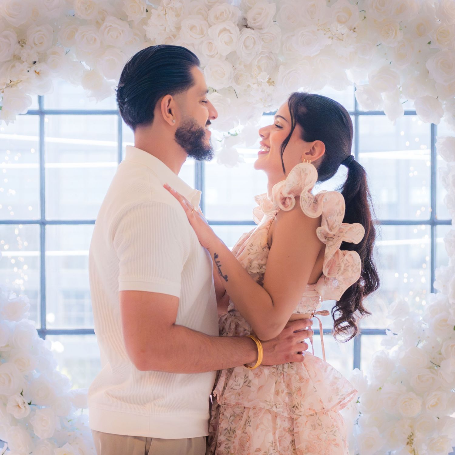 Couple embracing and smiling at each other in front of white floral heart arch.