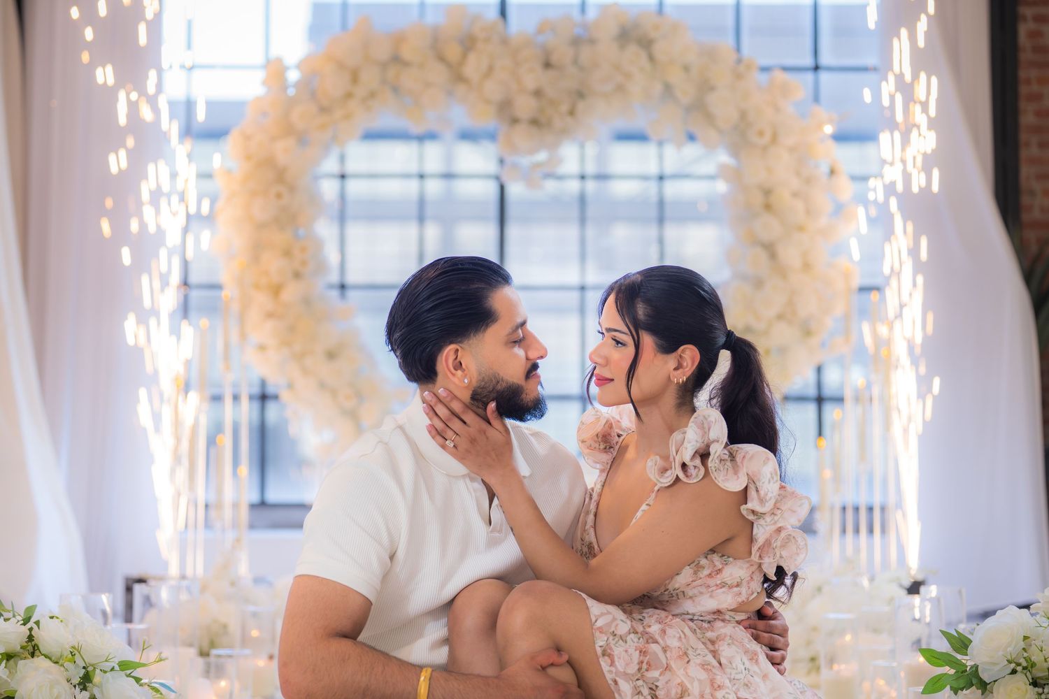 Couple gazing at each other under heart-shaped white floral arch with sparklers.