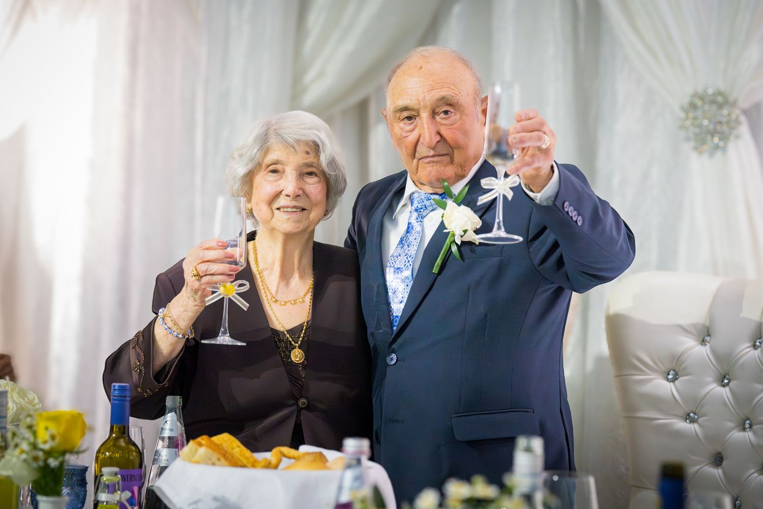 Elderly couple raising champagne glasses in a toast at their anniversary celebration with floral decorations behind them.