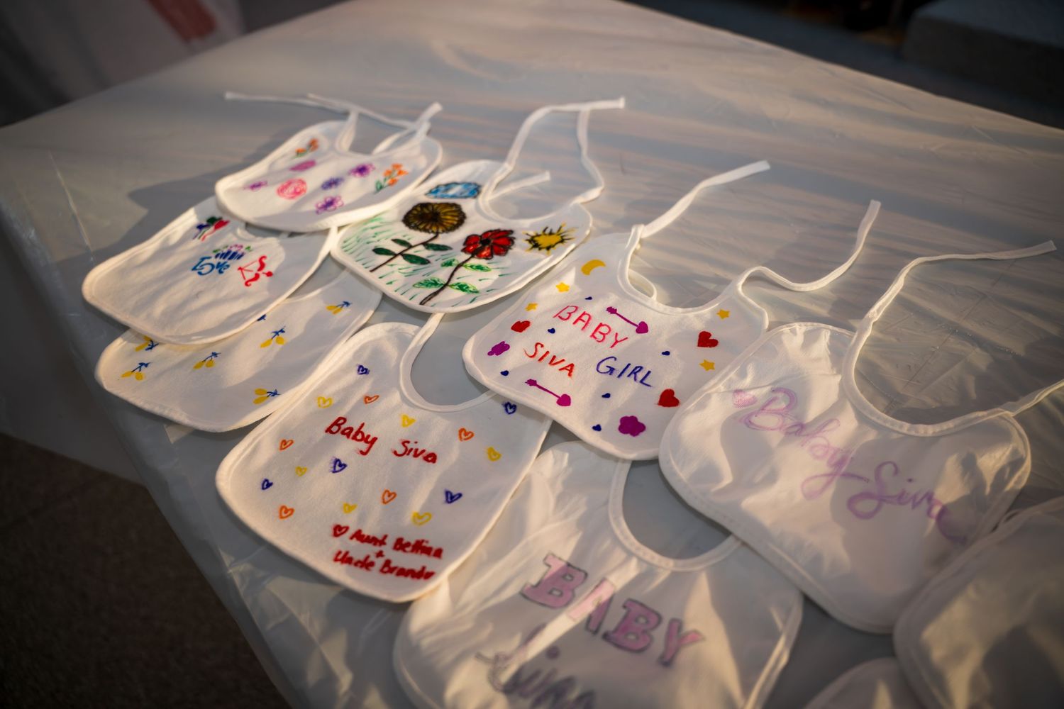 Collection of white baby bibs decorated with colorful designs and messages laid out on a table.