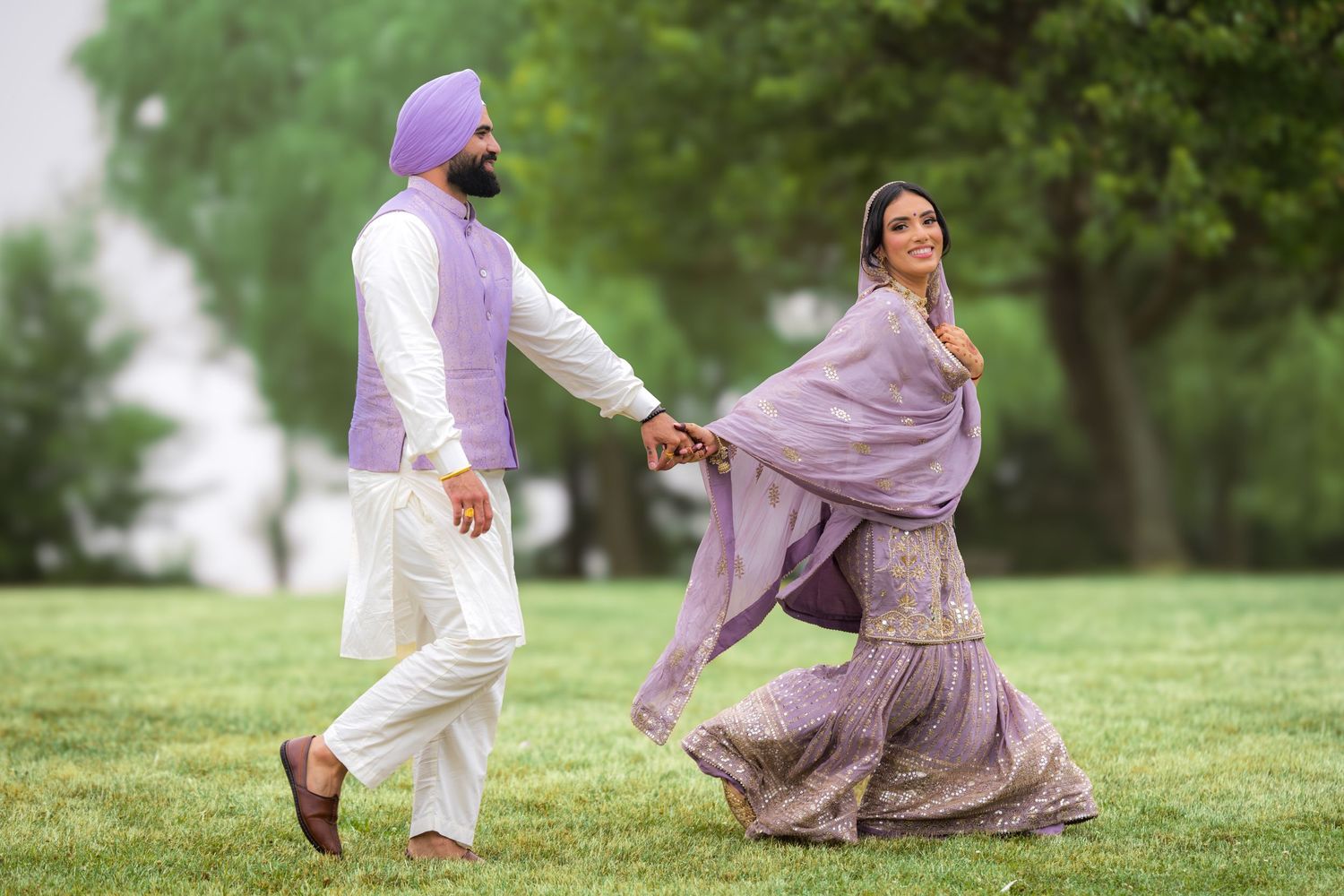Playful couple runs up a grassy hill in coordinated purple and white traditional outfits.