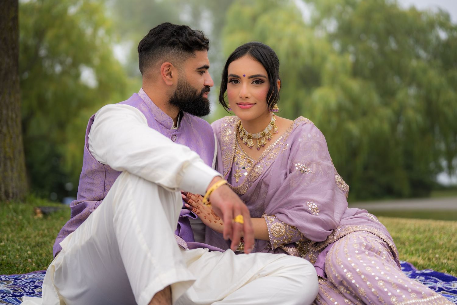 Couple in coordinated lavender and white outfits sitting together on grass during a photoshoot.