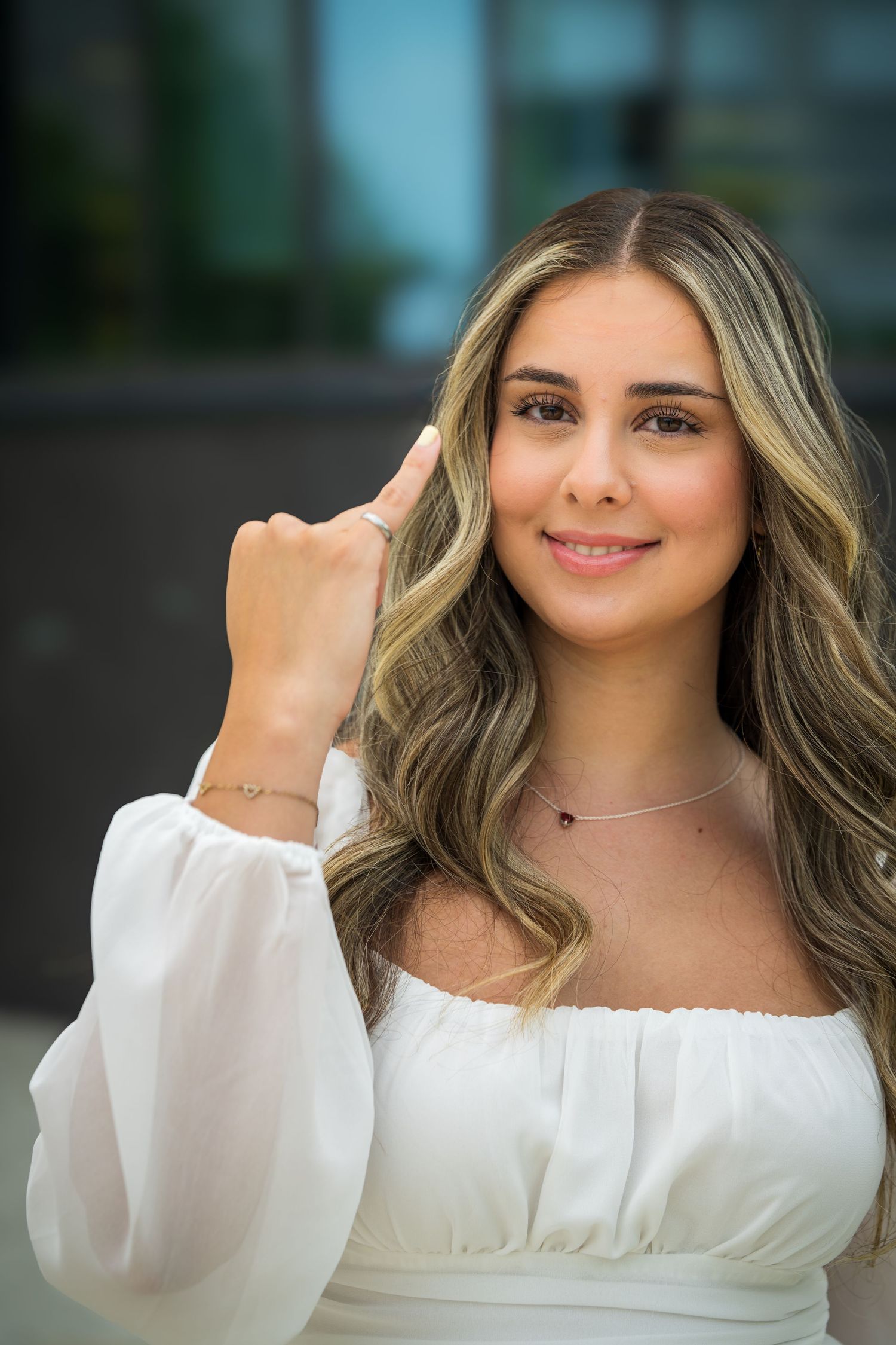 Graduate in white off-shoulder dress with wavy blonde hair poses elegantly against blurred background.