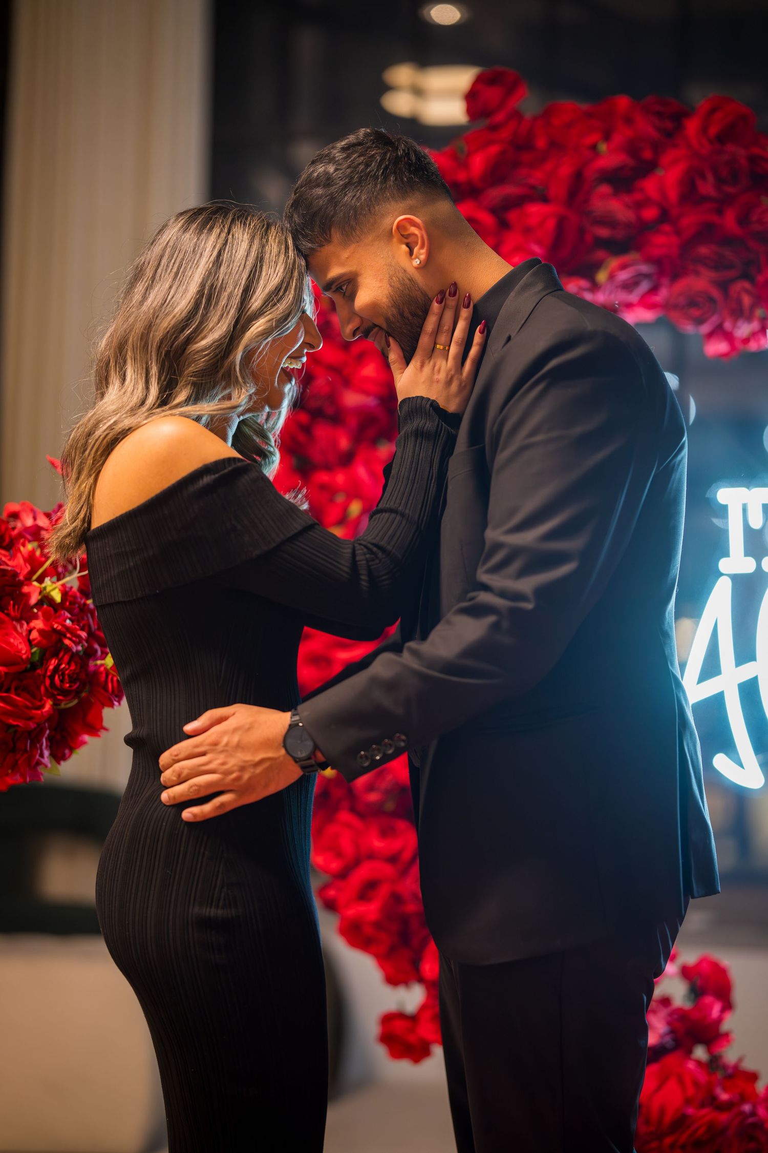 Couple embracing during proposal with red rose backdrop and blue neon lighting.