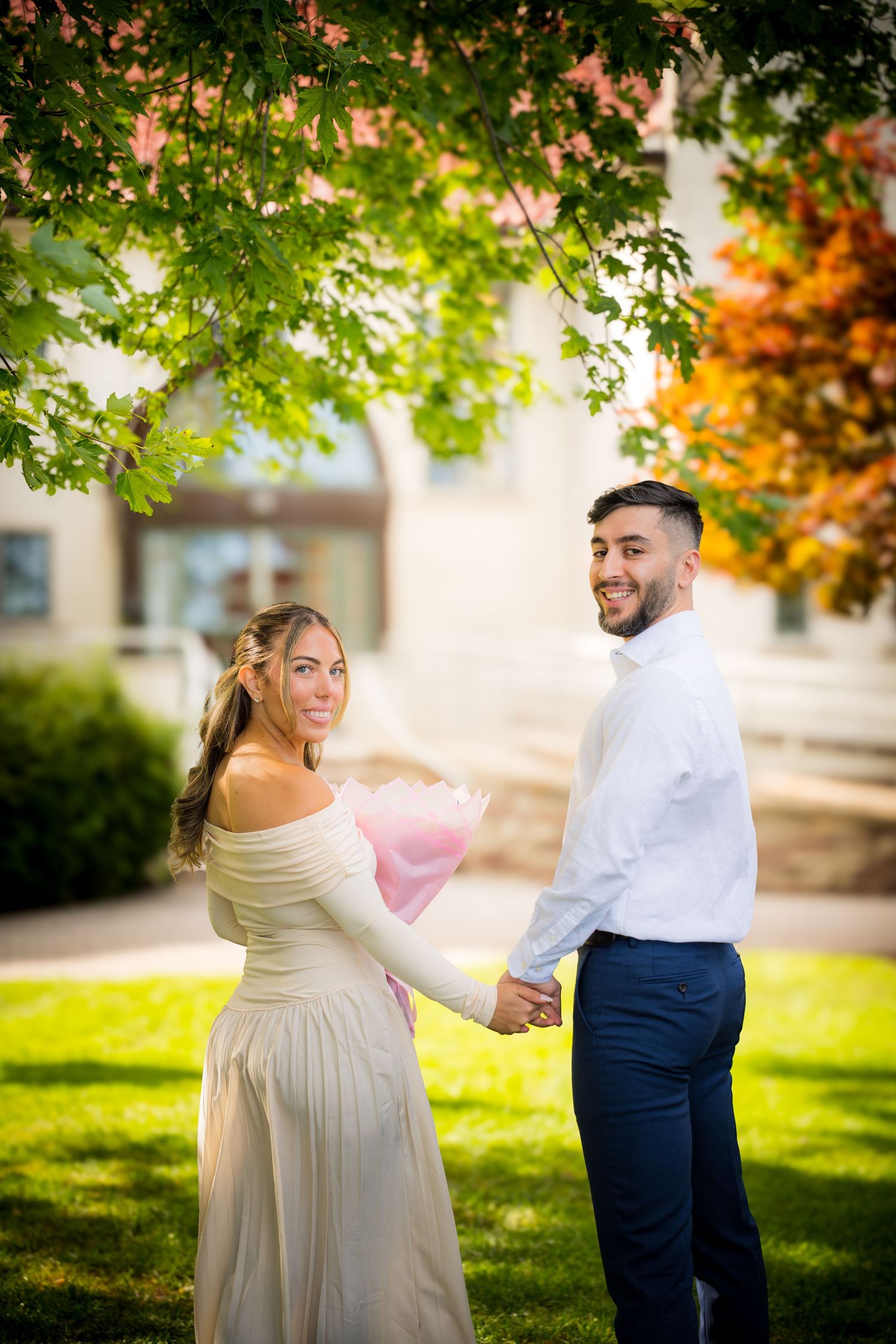 Couple holding hands outdoors, woman in beige gown with pink bouquet, man in white shirt, surrounded by colorful autumn trees.