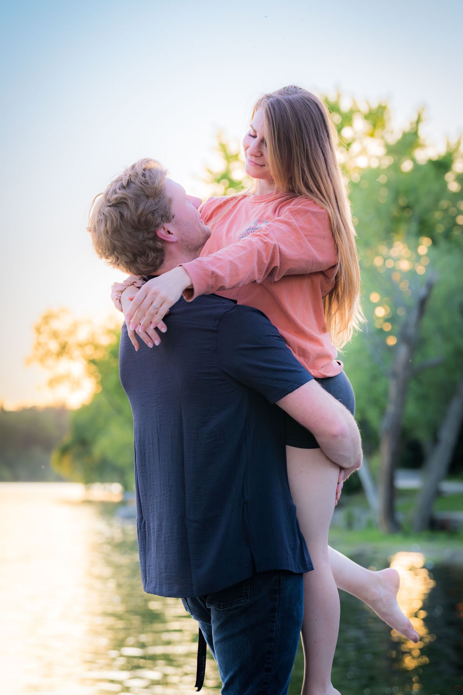 Man lifting a smiling woman in his arms near a lake at golden hour with trees in background.