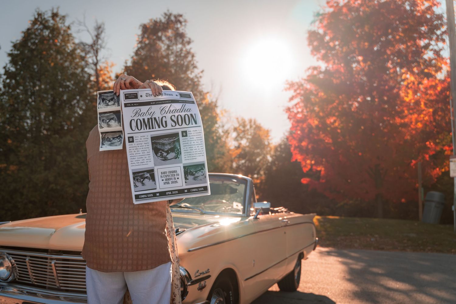 Person holding a newspaper with Coming Soon headline beside a vintage convertible car in autumn sunlight.