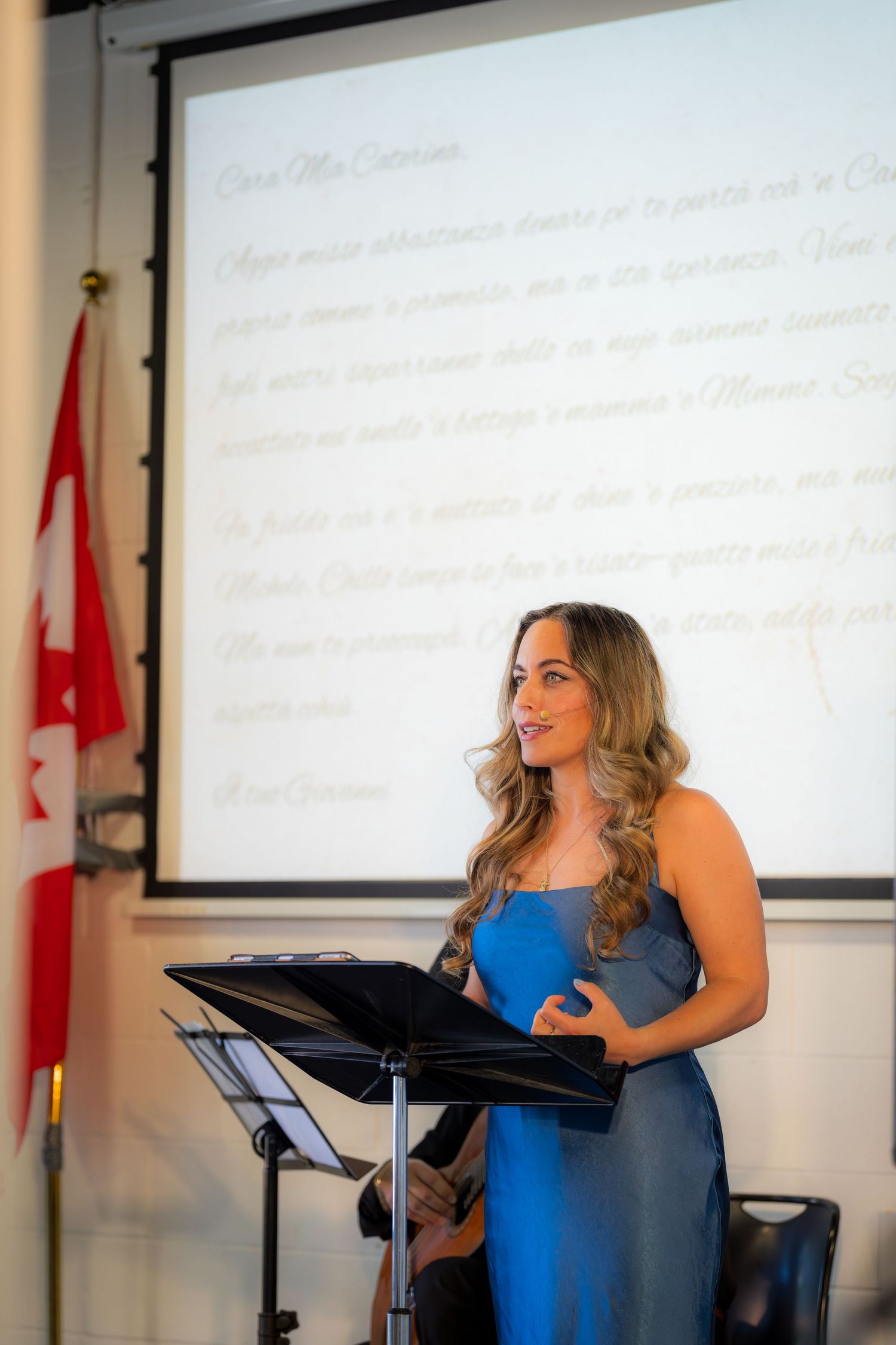 Woman in blue dress speaking at podium with Italian text projected on screen behind her.