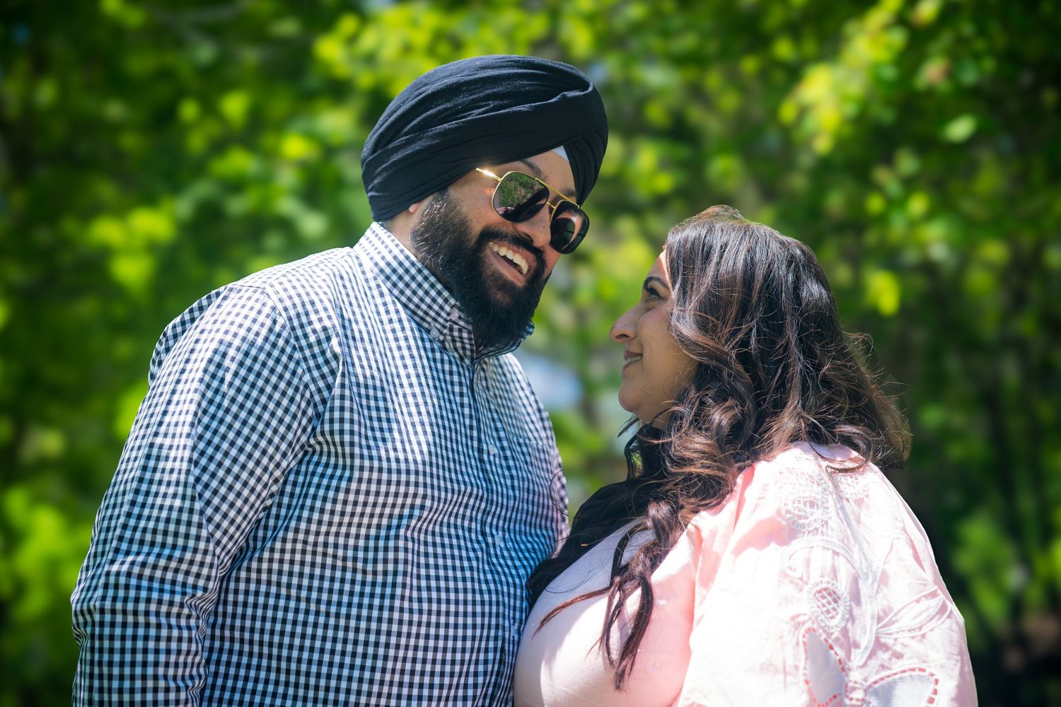 A Sikh man in a turban and sunglasses shares a joyful moment with a woman in a pink dress outdoors.
