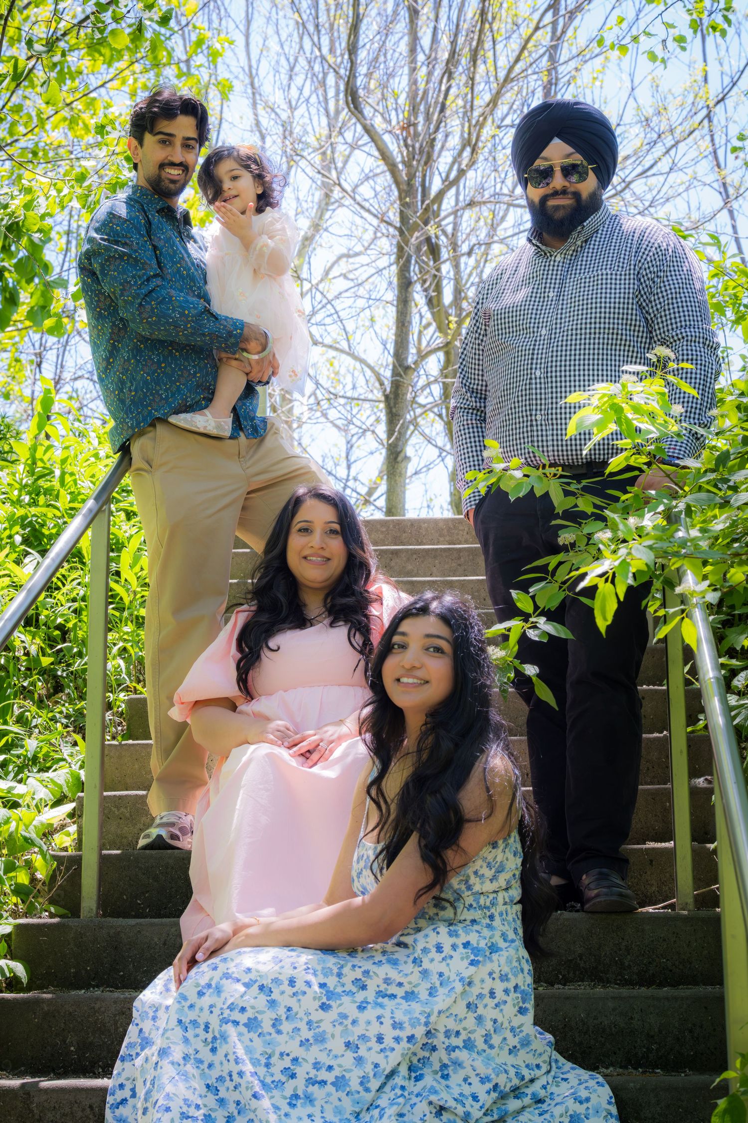 A South Asian family of four poses on outdoor steps surrounded by bright green foliage.