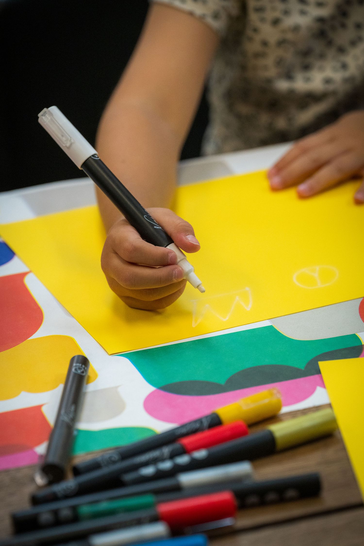 Child drawing on yellow paper with a white marker surrounded by colorful markers on a table.