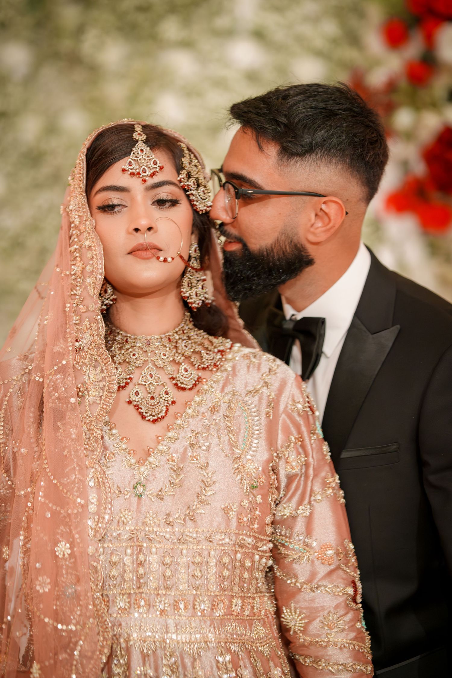 Couple in traditional South Asian wedding attire sharing intimate moments against floral backdrop.