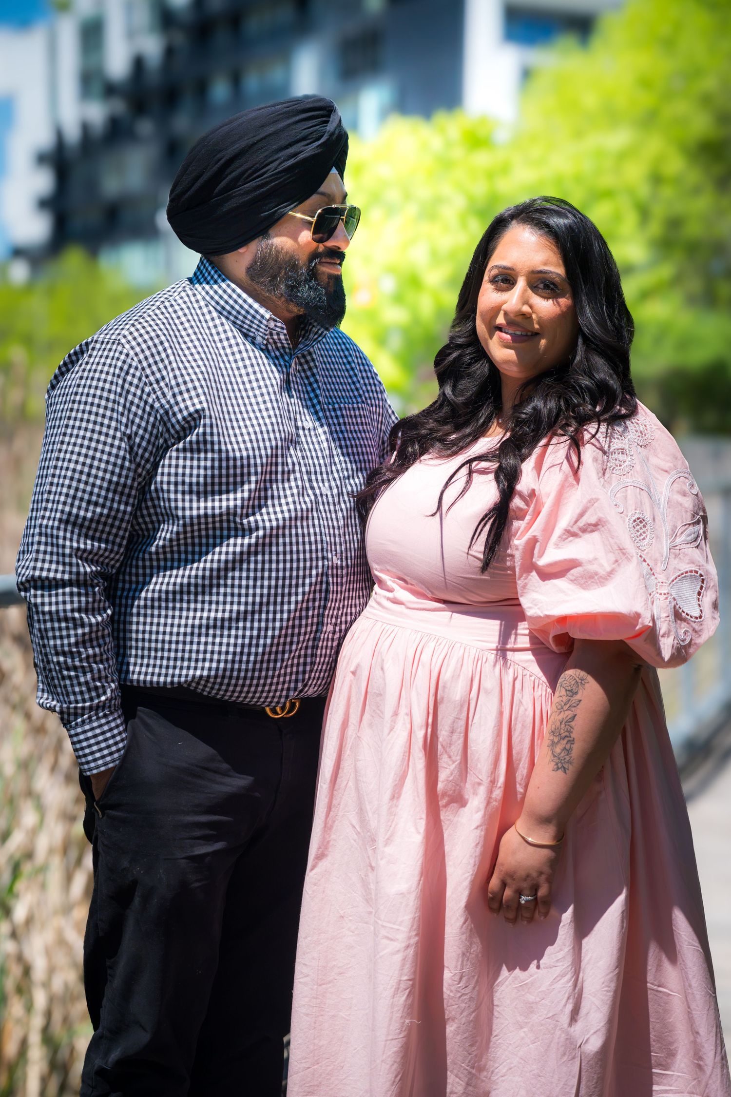 A Sikh man in a turban and checkered shirt stands with a woman in a pink dress outdoors.