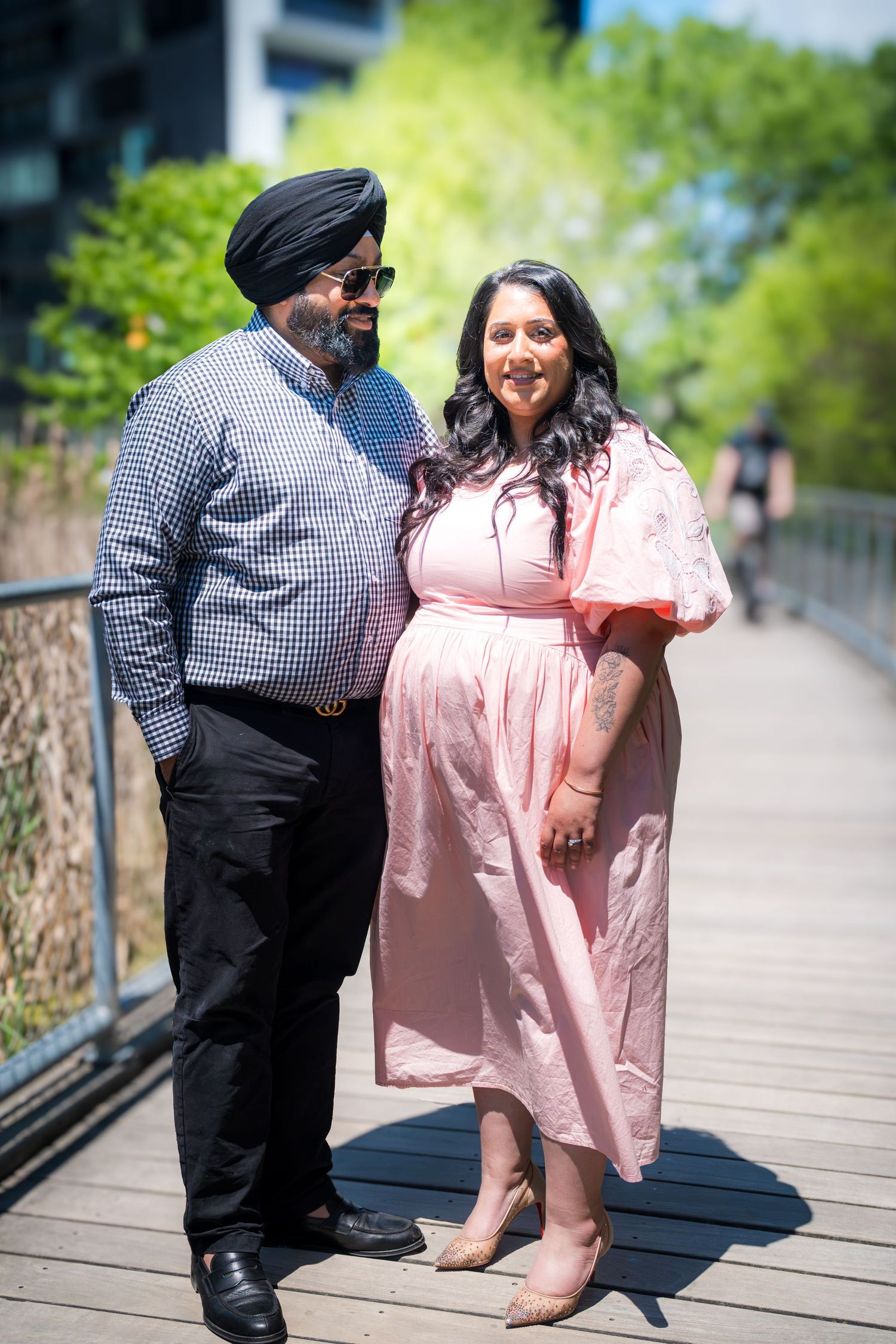 A Sikh couple poses on a sunny bridge, man in turban and checkered shirt, woman in pink dress.