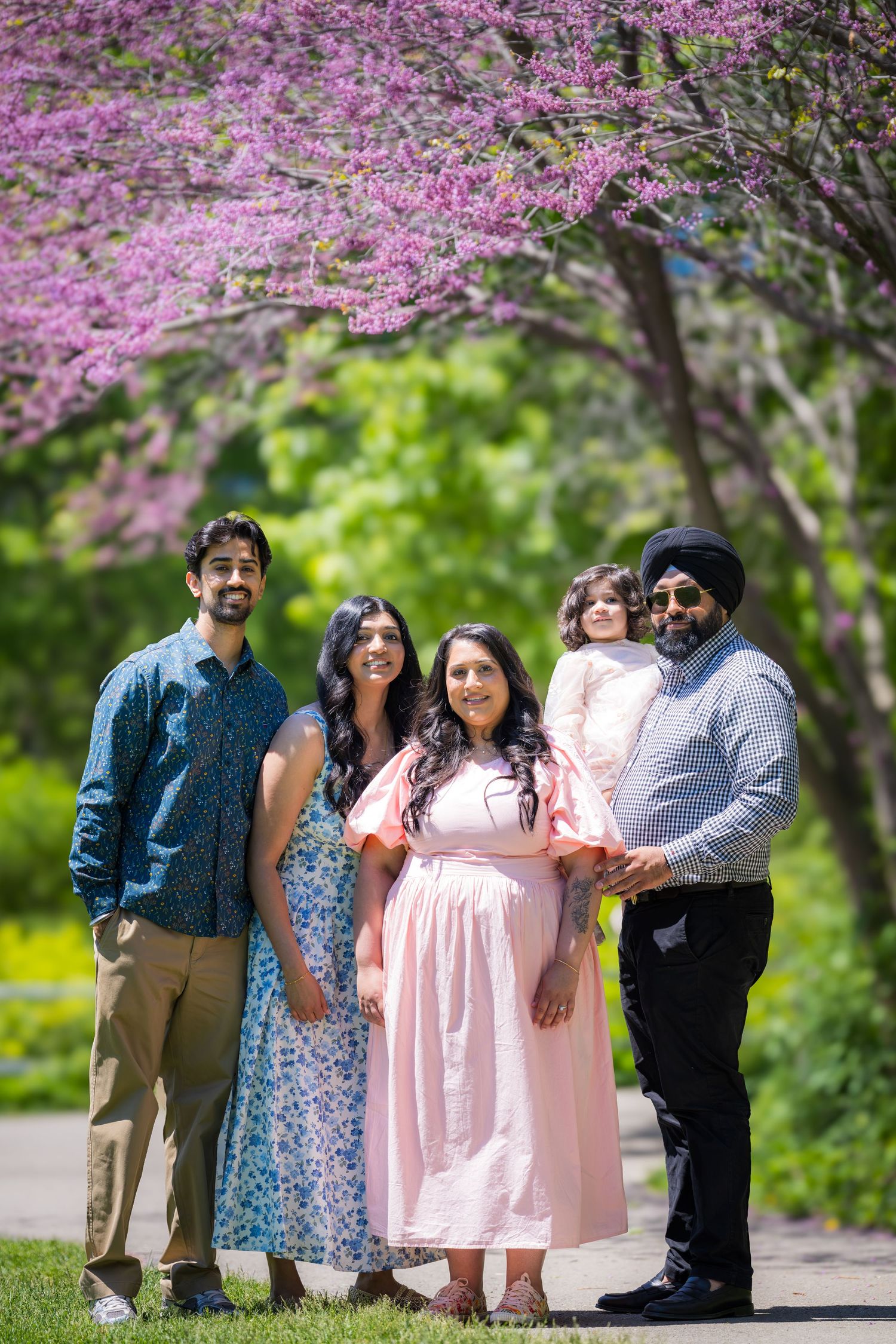 A Sikh family of five poses under blooming pink redbud tree, father in turban holding child in spring park.