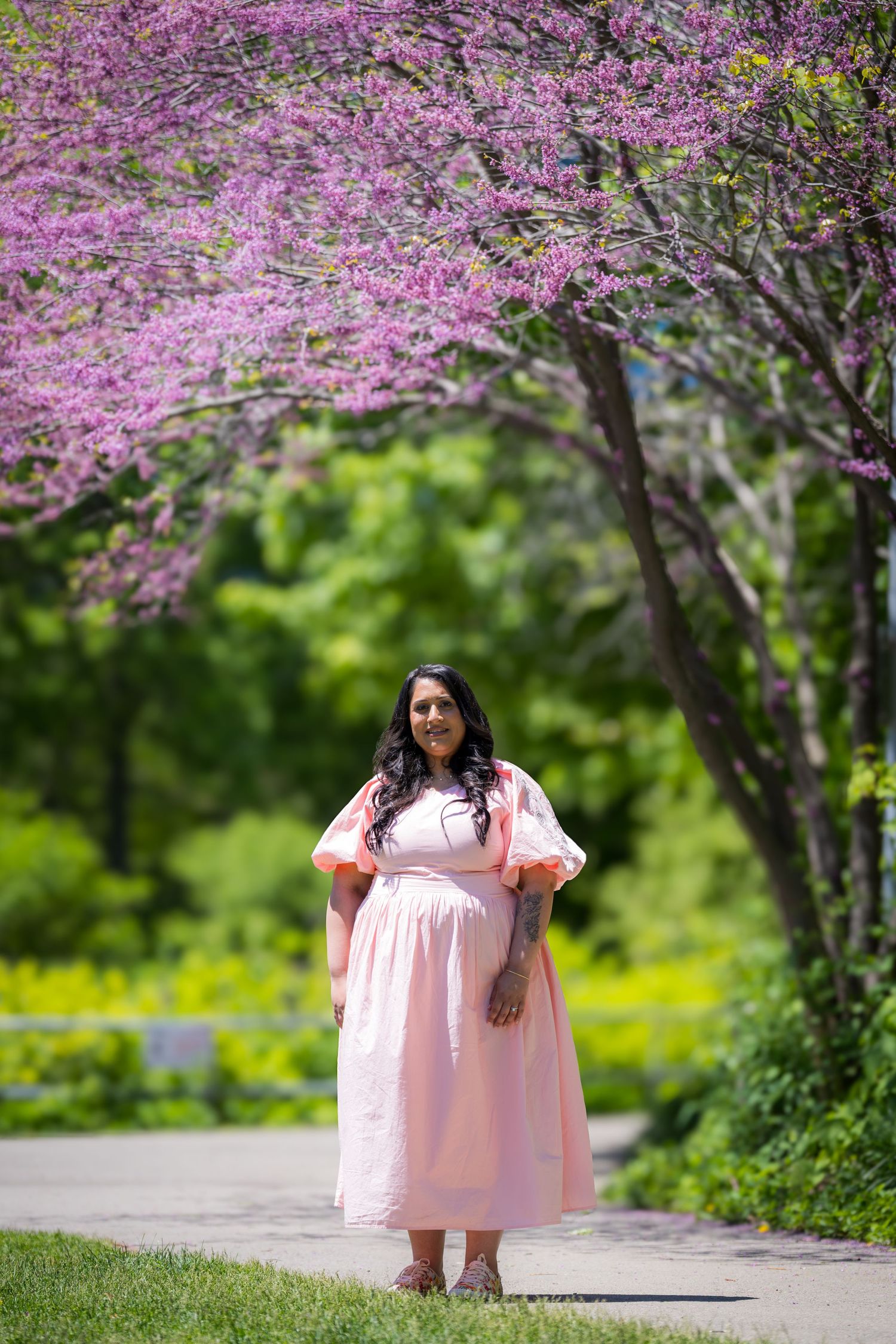 Pregnant South Asian woman in pink maxi dress standing under blooming pink redbud trees.