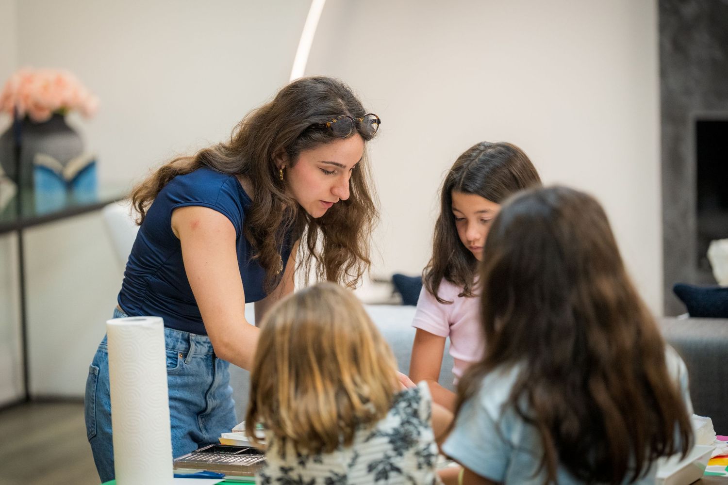 A teacher leans over a table helping young girls with an art activity in a bright modern classroom.