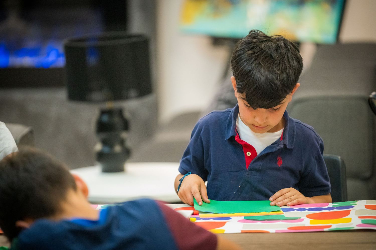 A young boy in a navy polo shirt focuses on a colorful paper craft activity at a table.