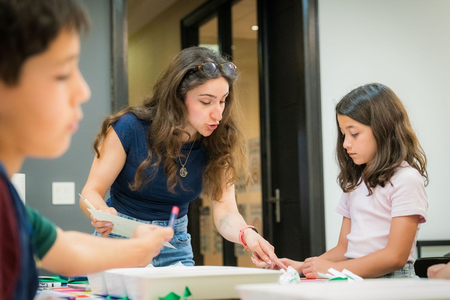 A teacher assists a young girl with a colorful hands-on art project at a table in a bright classroom.