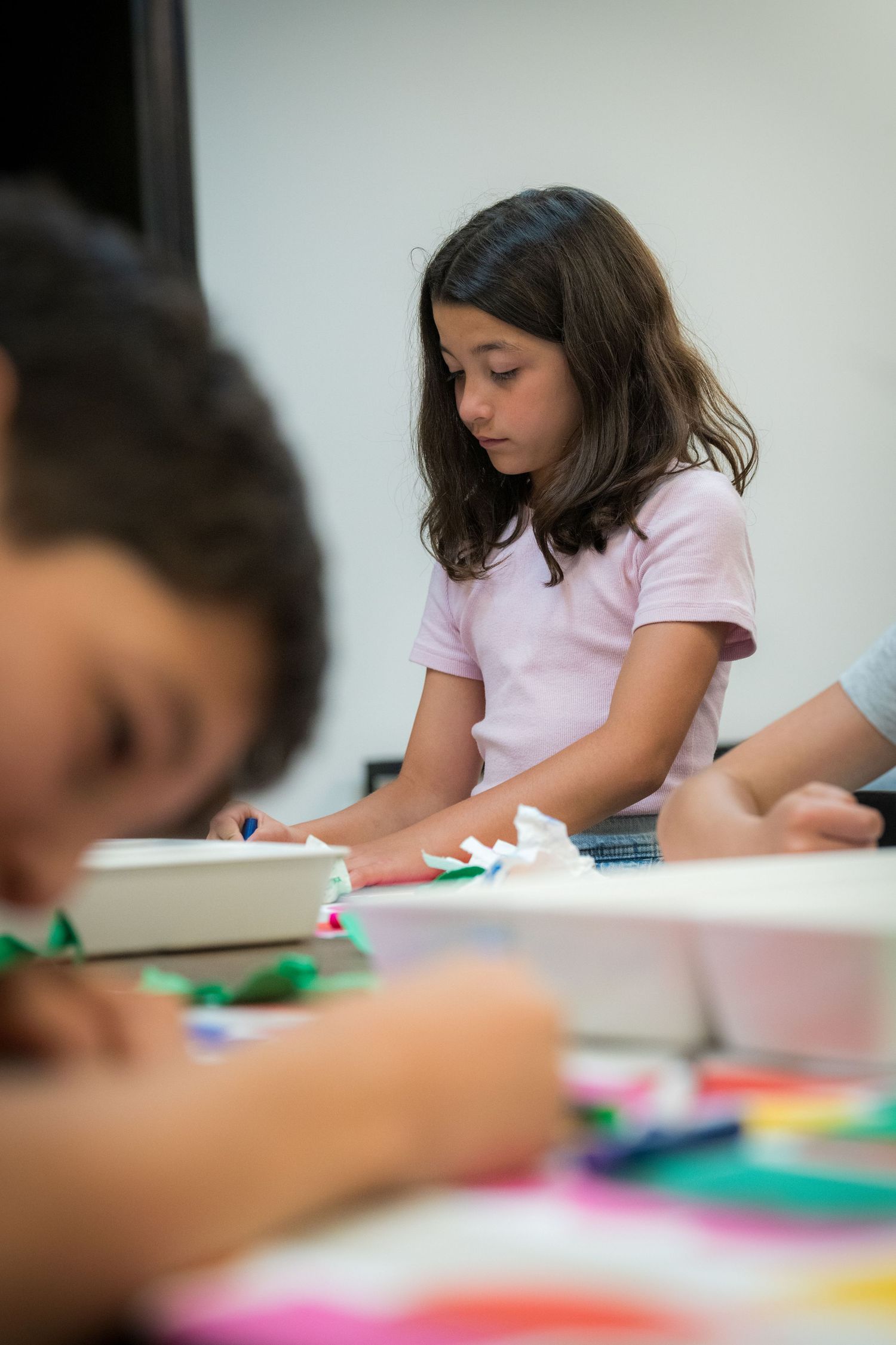 A young girl in a pink shirt concentrates on a colorful paper art project at a classroom table.