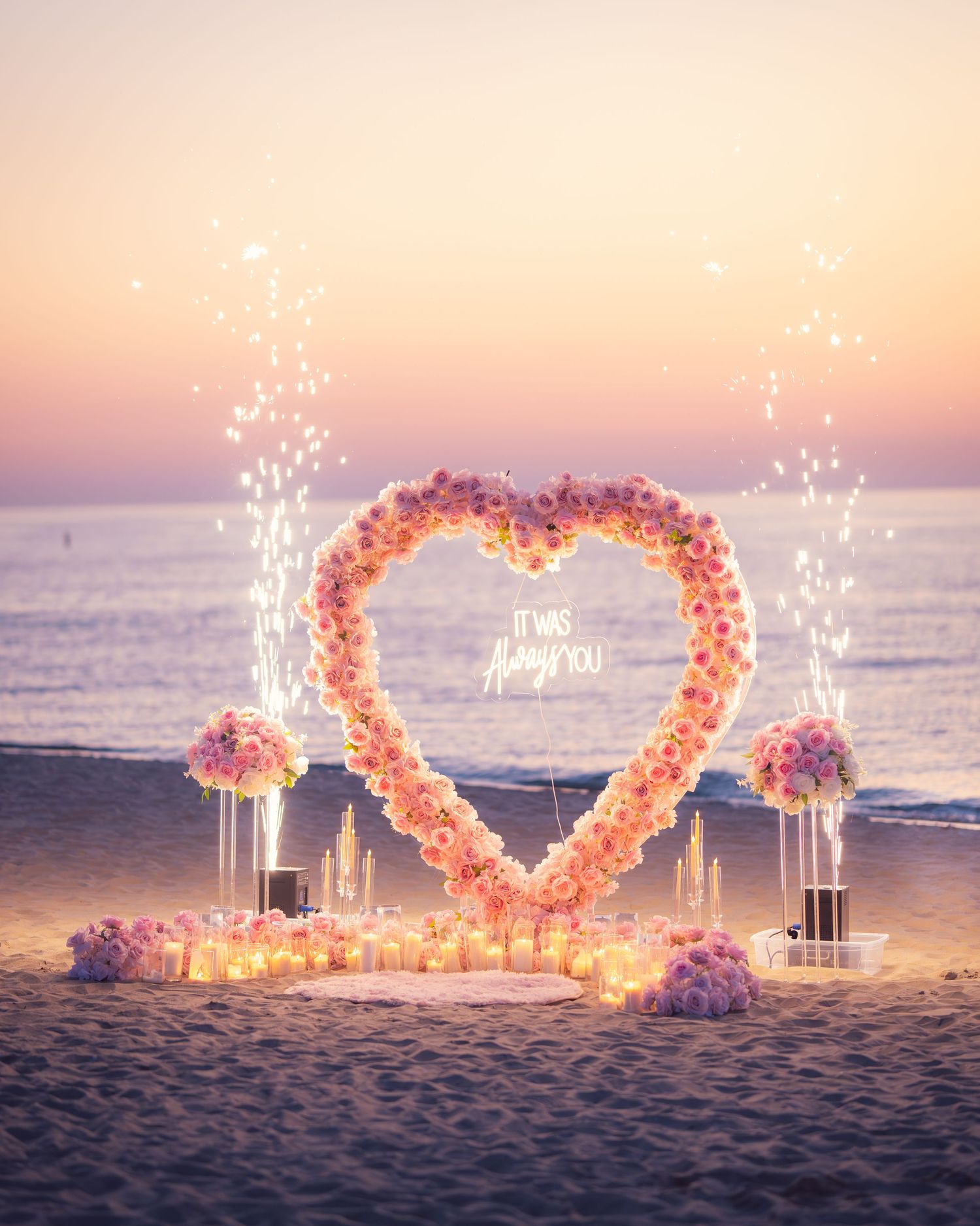 A romantic heart-shaped floral arch with sparklers and candles glows at sunset on a peaceful beach.