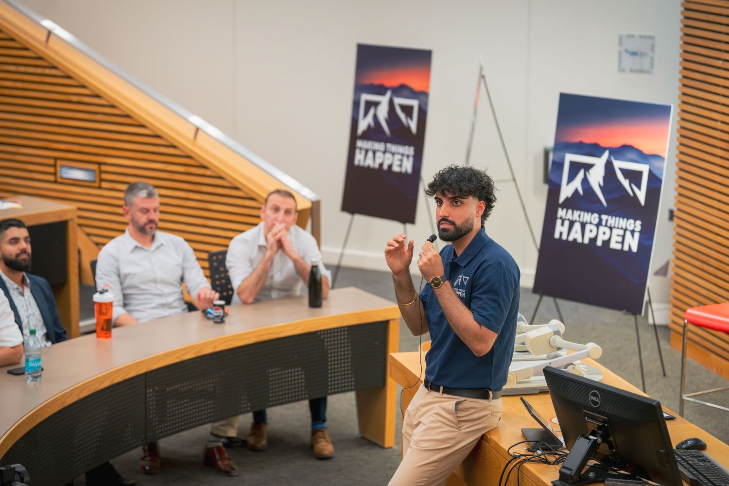 A presenter in a navy polo shirt leads a discussion in a modern classroom setting with tiered seating.