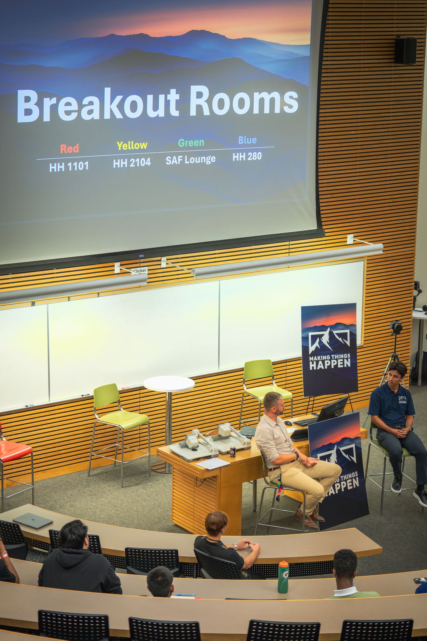 Lecture hall with breakout room assignments projected and speakers on stage.