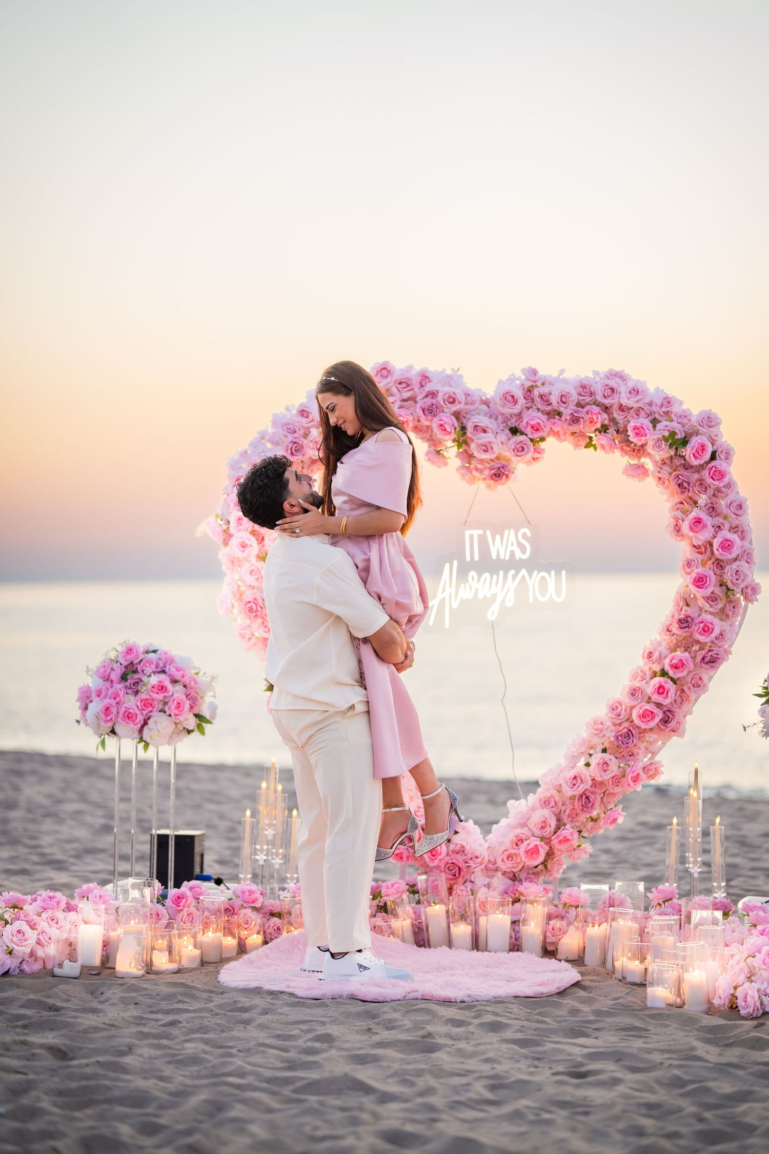Romantic beach proposal setup with heart-shaped pink floral arch and candles at sunset creates magical moment.