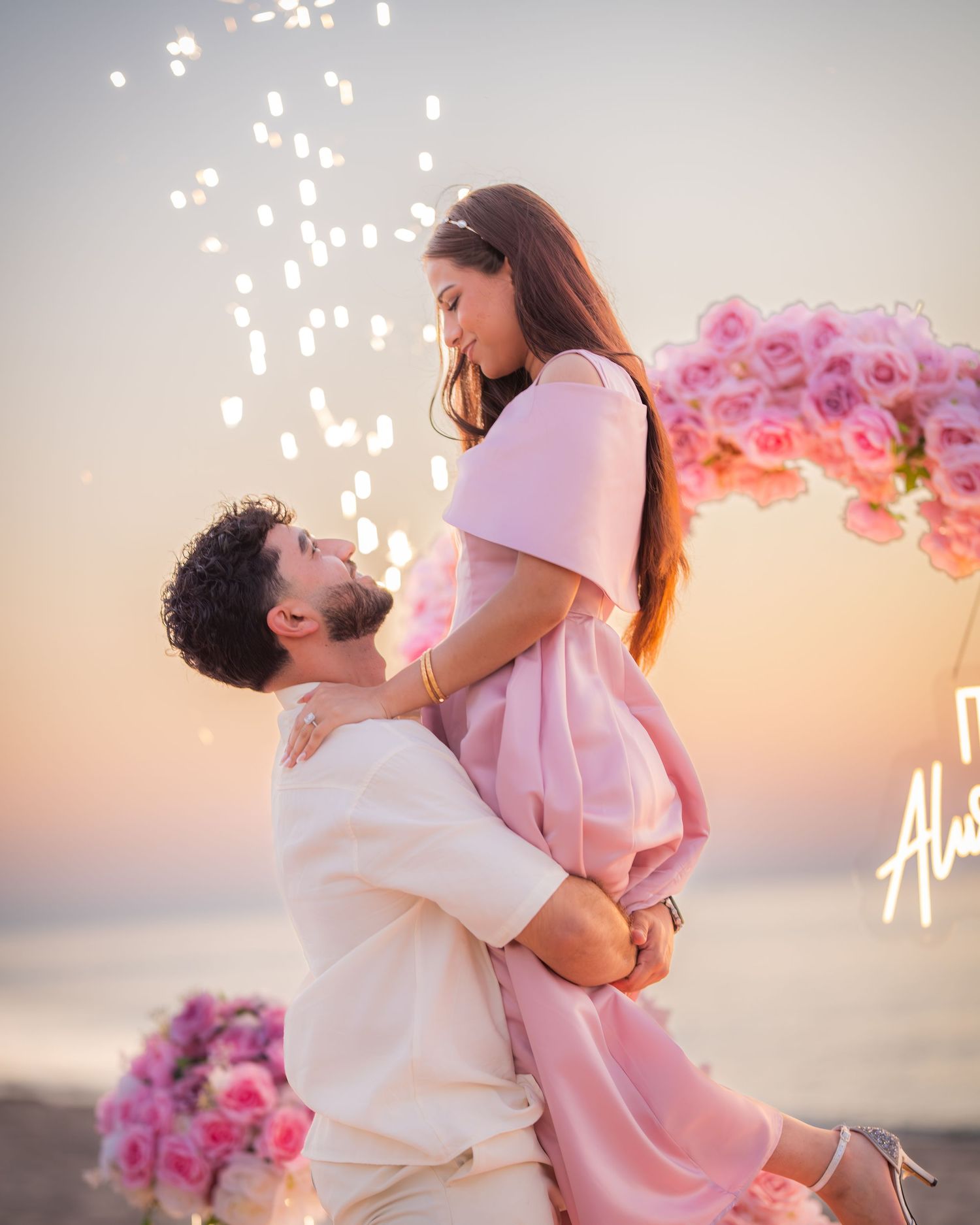 Sunset beach proposal moment under pink floral heart arch with sparkling lights.