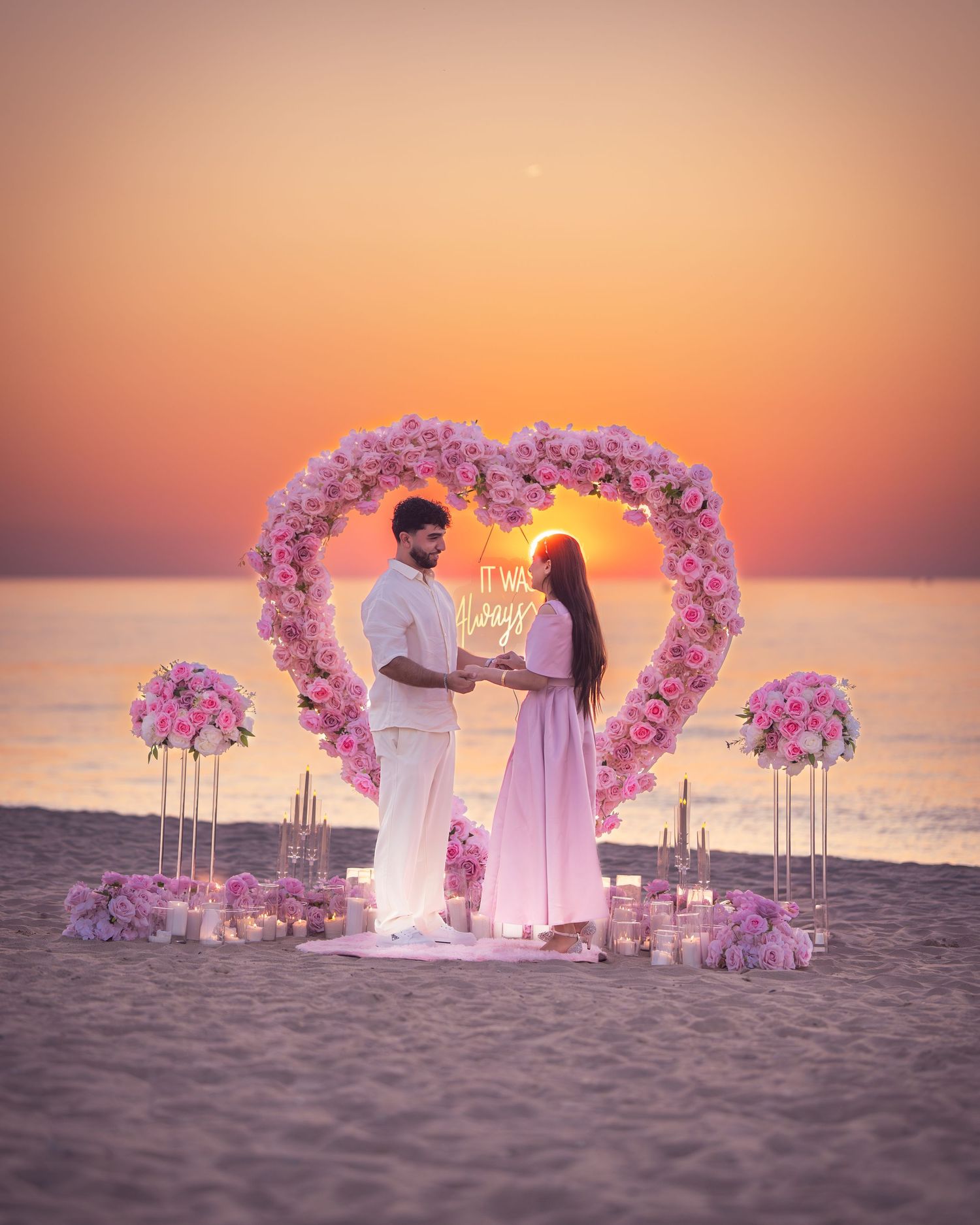 A romantic beach proposal setup with a heart-shaped floral arch and pink decorations at sunset.