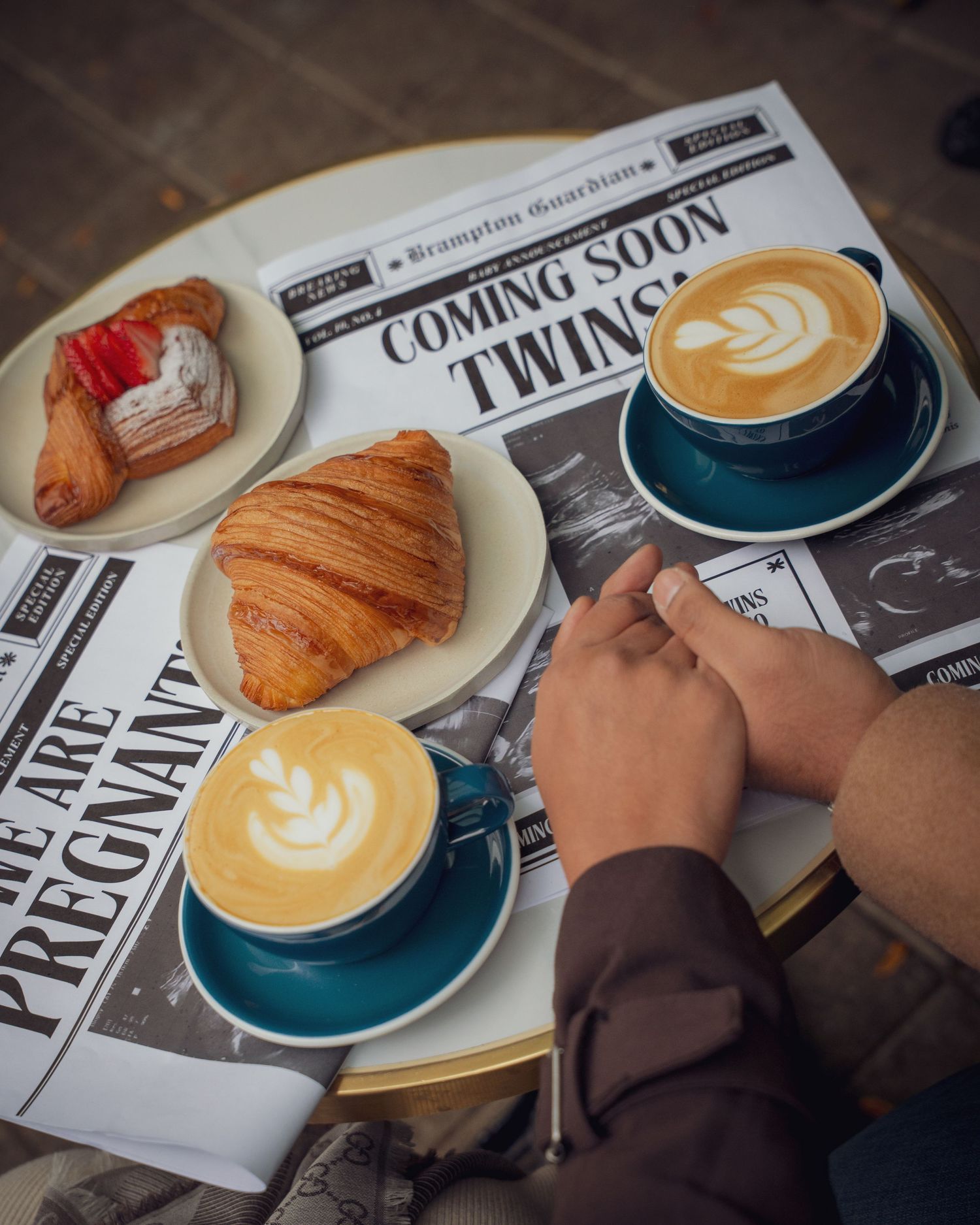 Breakfast scene with two lattes, a croissant, and pastry on a newspaper with 'Coming Soon Twins' headline.