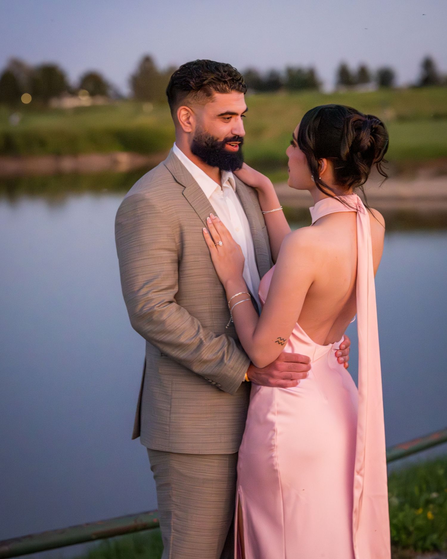 Couple shares intimate moment by lakeside during golden hour wearing pink dress and grey suit.