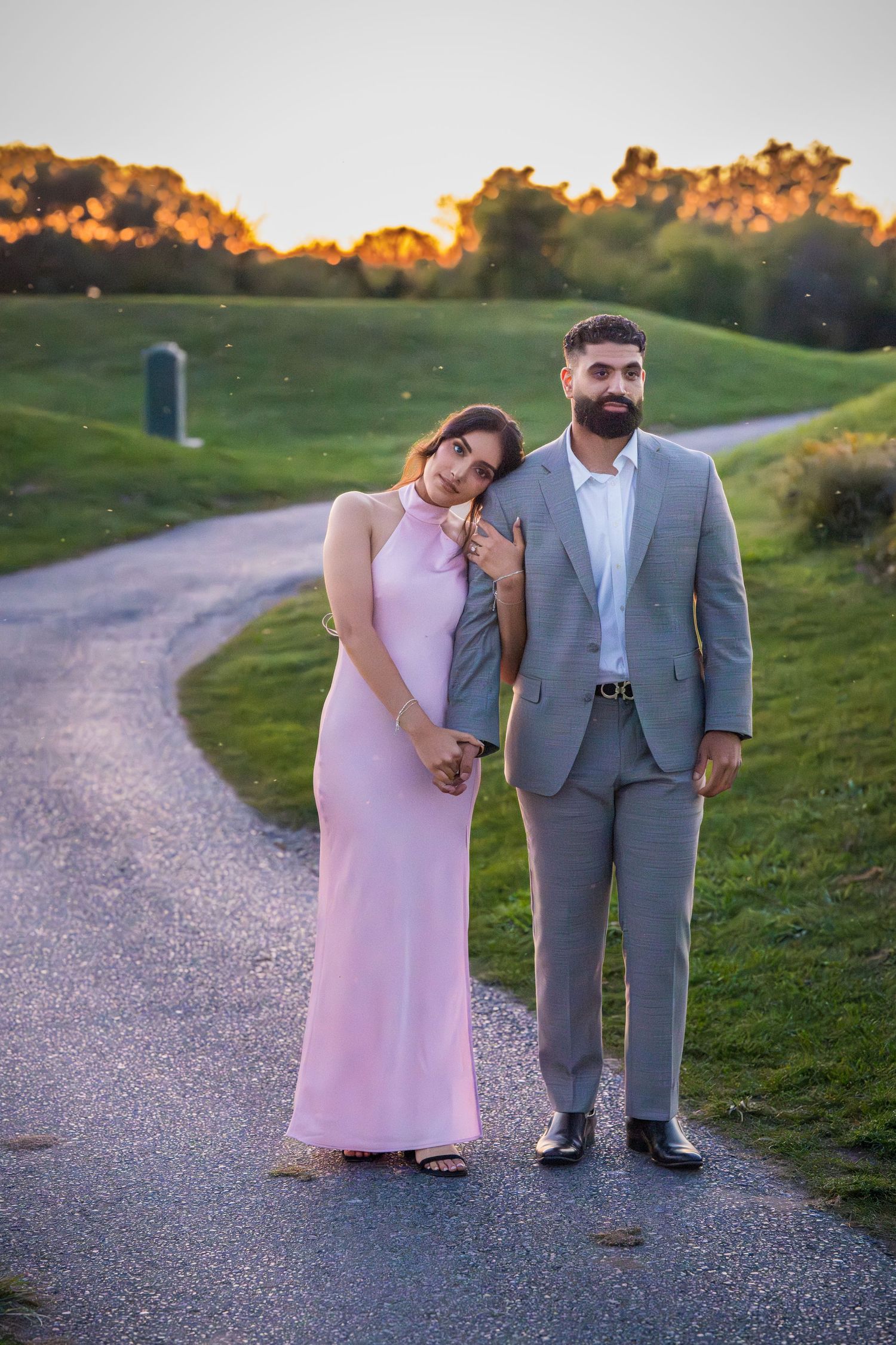 Two people pose playfully on a gravel path during sunset wearing formal attire.