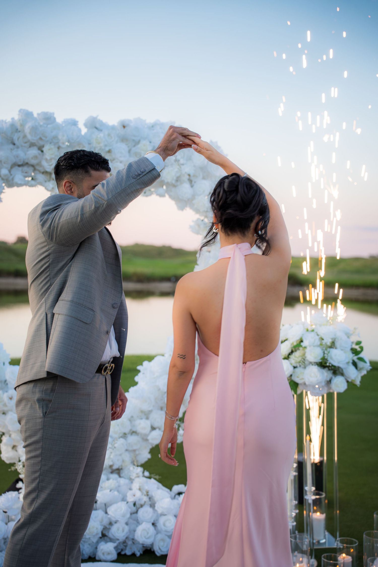 Couple dances under white floral arch with sparklers glowing in outdoor wedding setting.