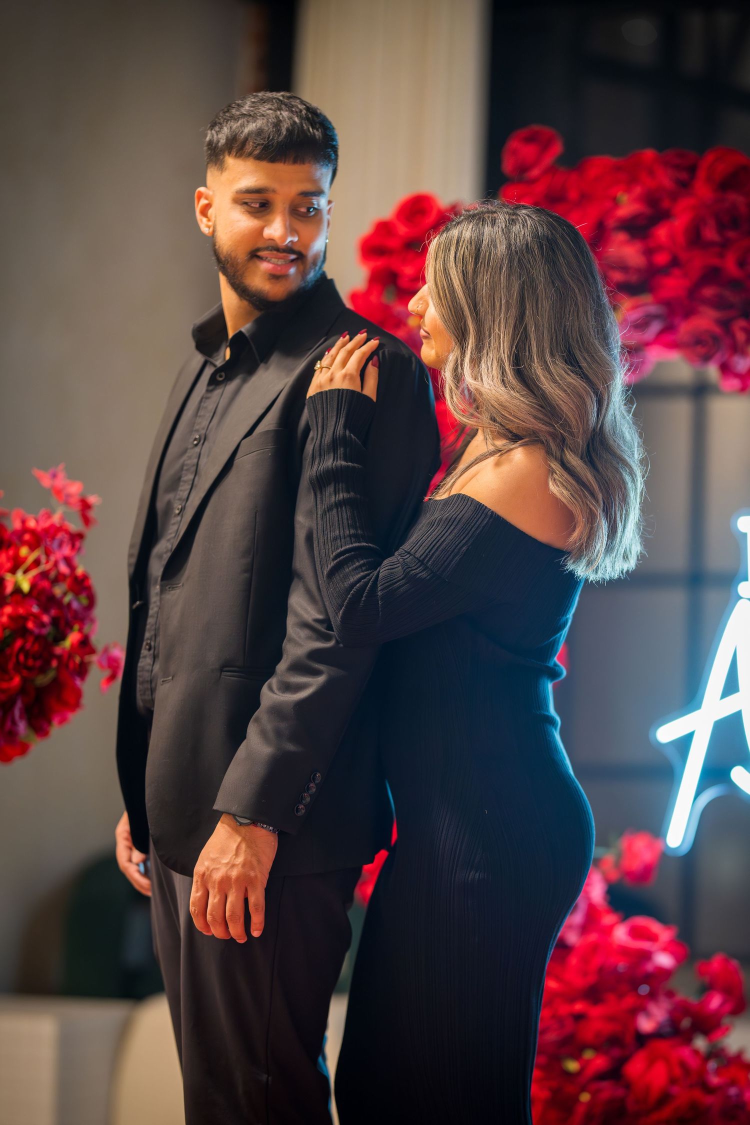 A couple embracing in formal attire against a backdrop of red roses and soft lighting.