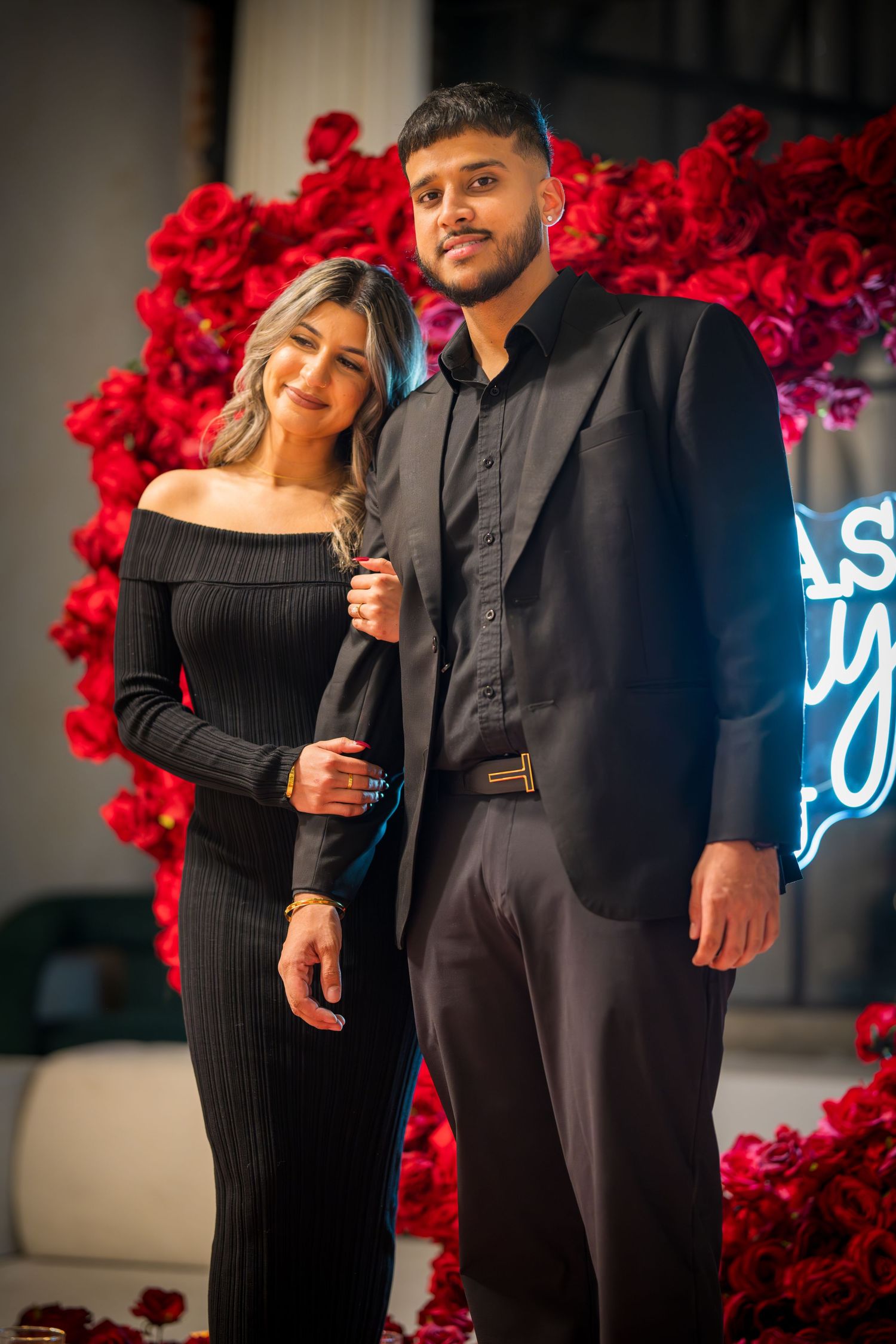 Stylish pair in matching black outfits pose before red rose wall and neon signage.