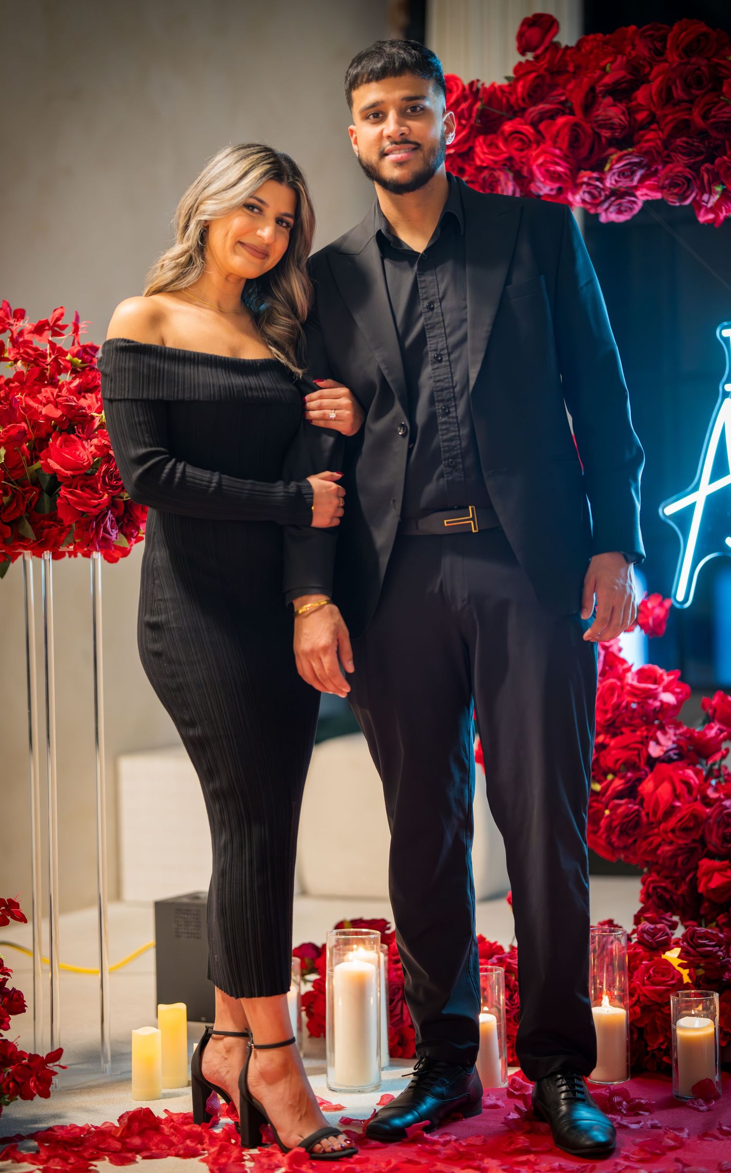 A couple poses in formal black attire against a backdrop of red roses and candles in a romantic setting.