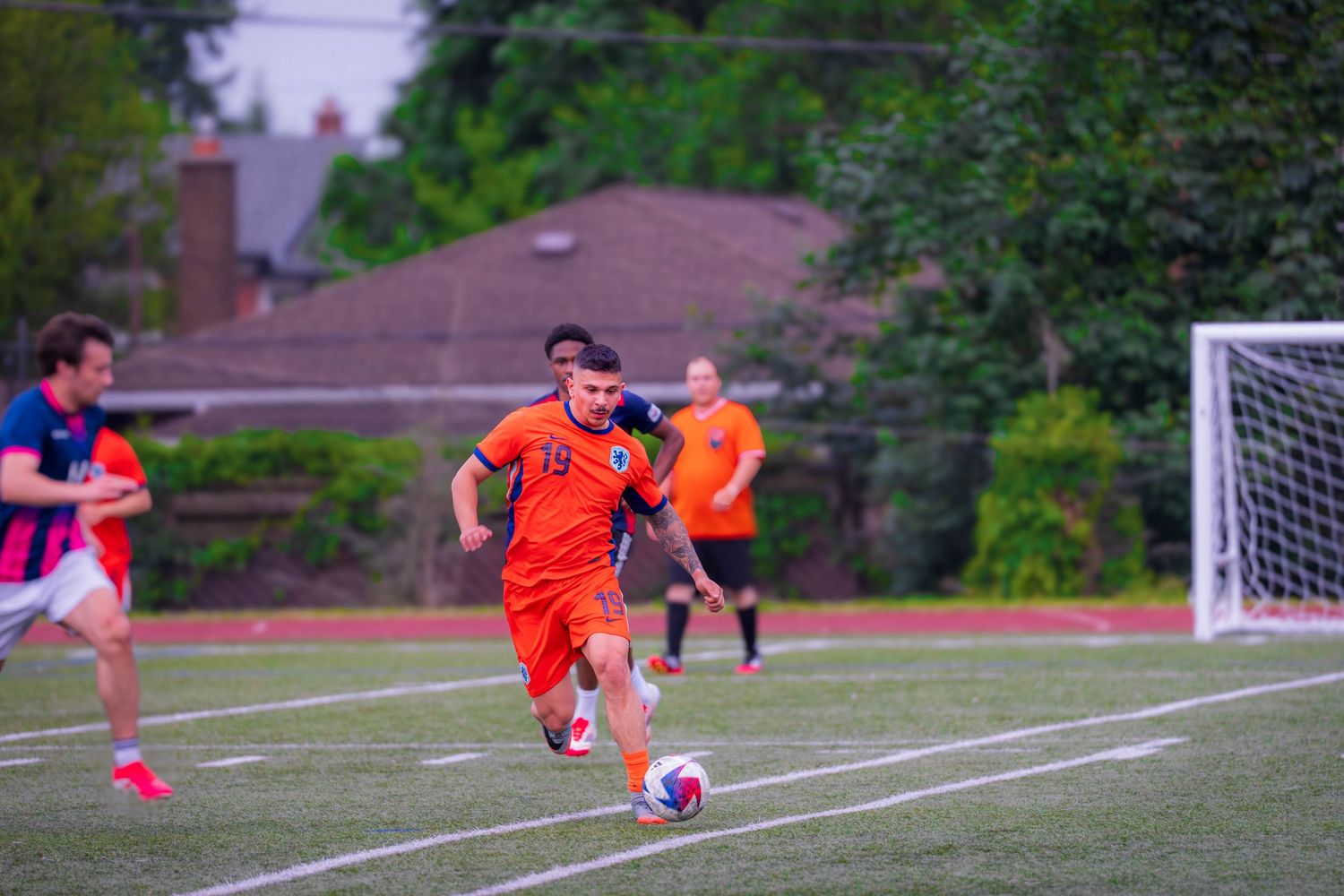 Soccer players in orange and navy uniforms battle for possession on a sports field.