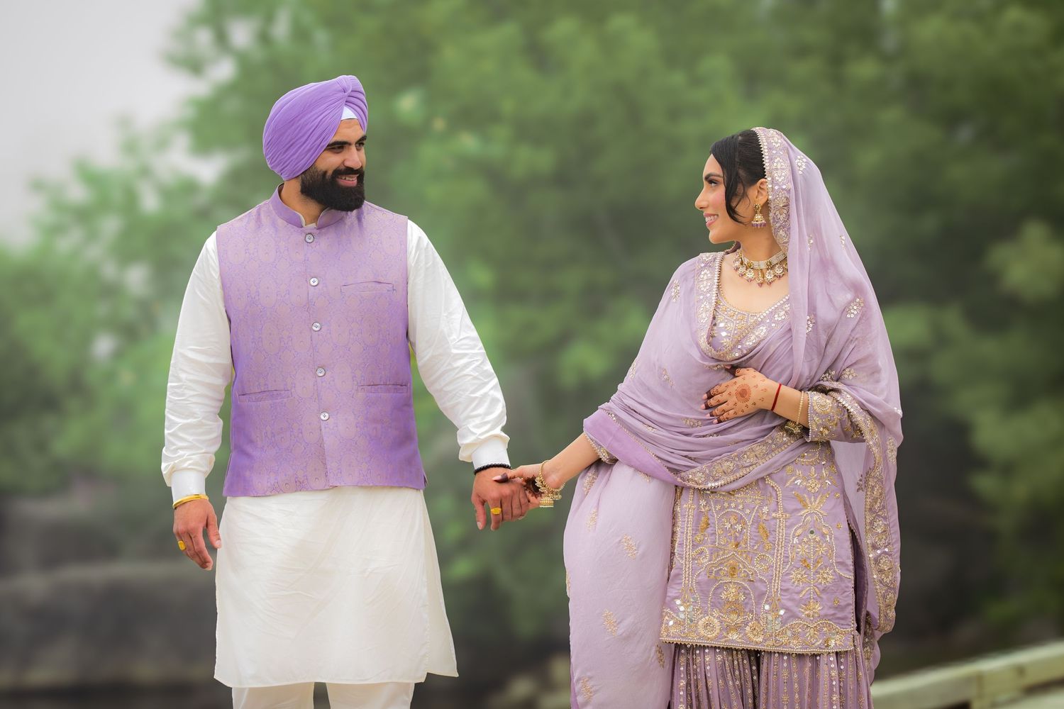 A couple in matching purple and white traditional outfits poses together outdoors.