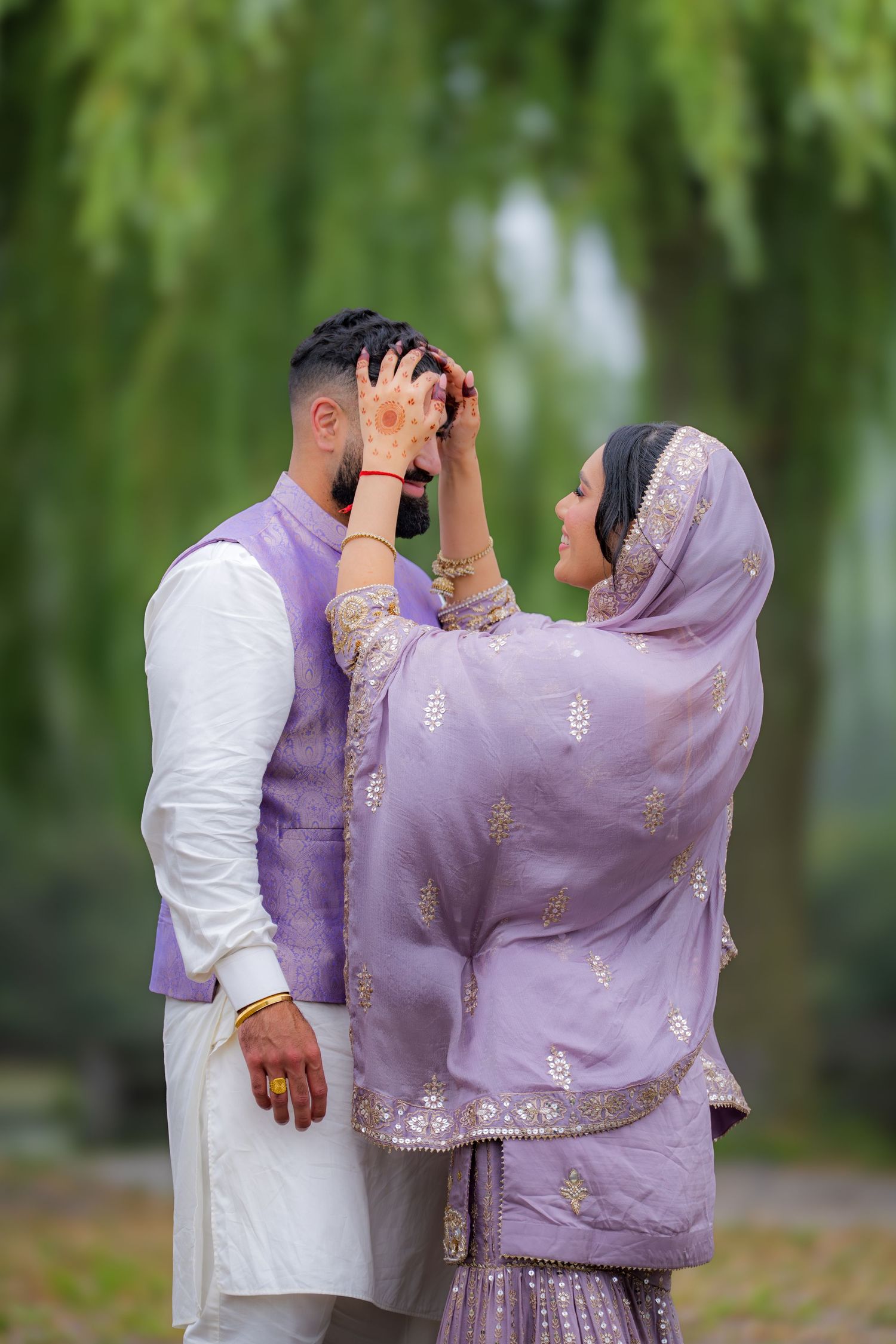 A couple in traditional Indian clothing poses playfully under willow trees in a park.