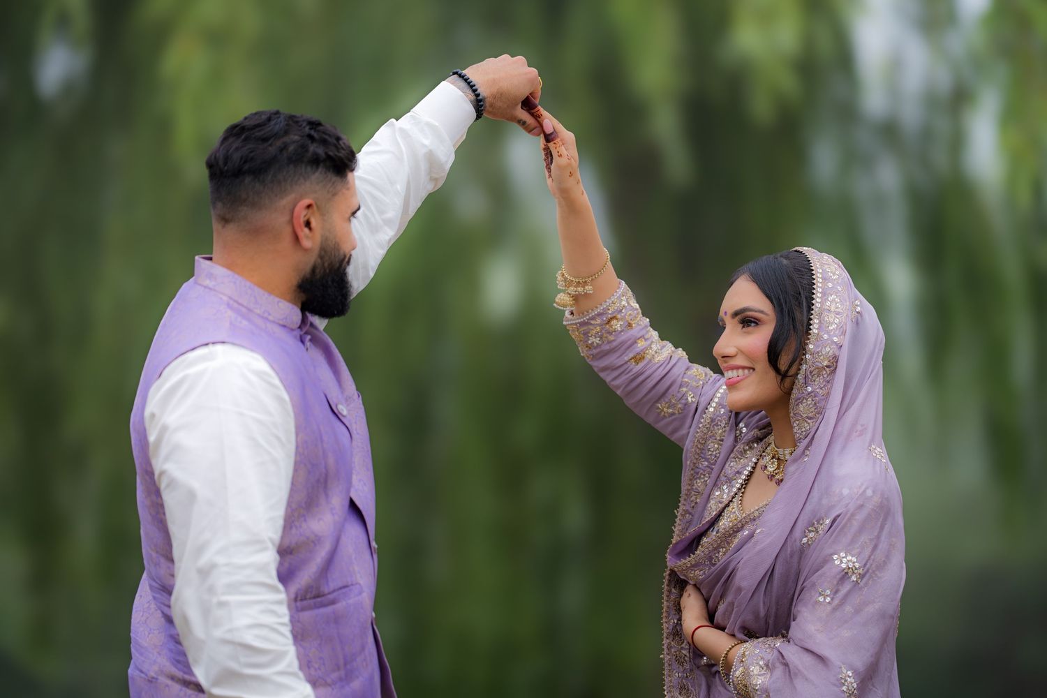 A couple in coordinating purple and white outfits dance together in a garden setting with willow trees.