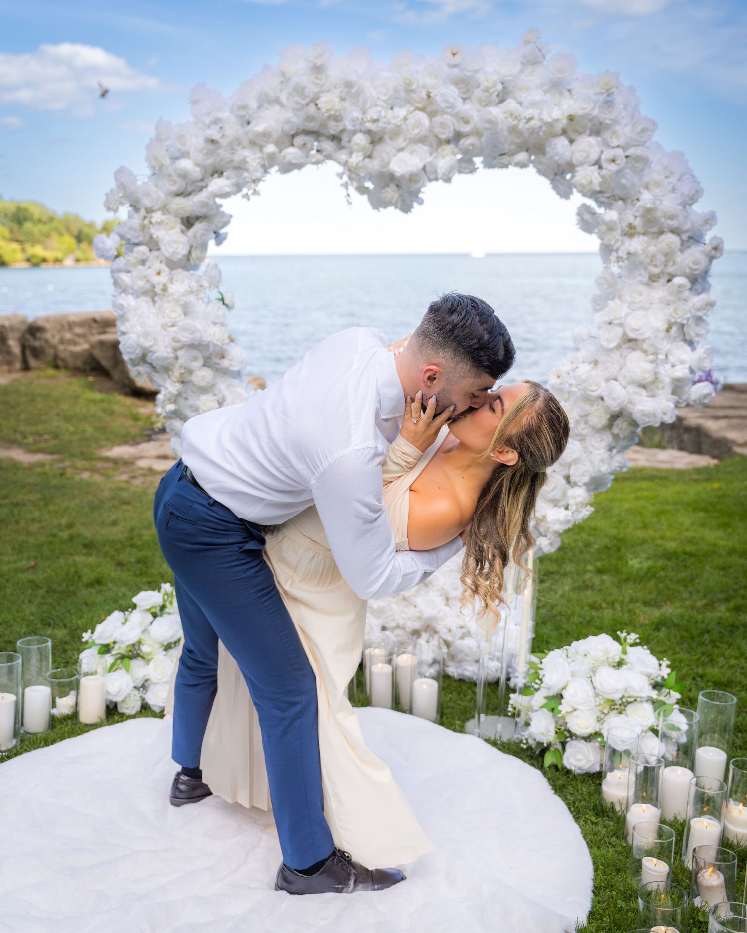 A romantic dip kiss under a white floral arch during a seaside proposal.